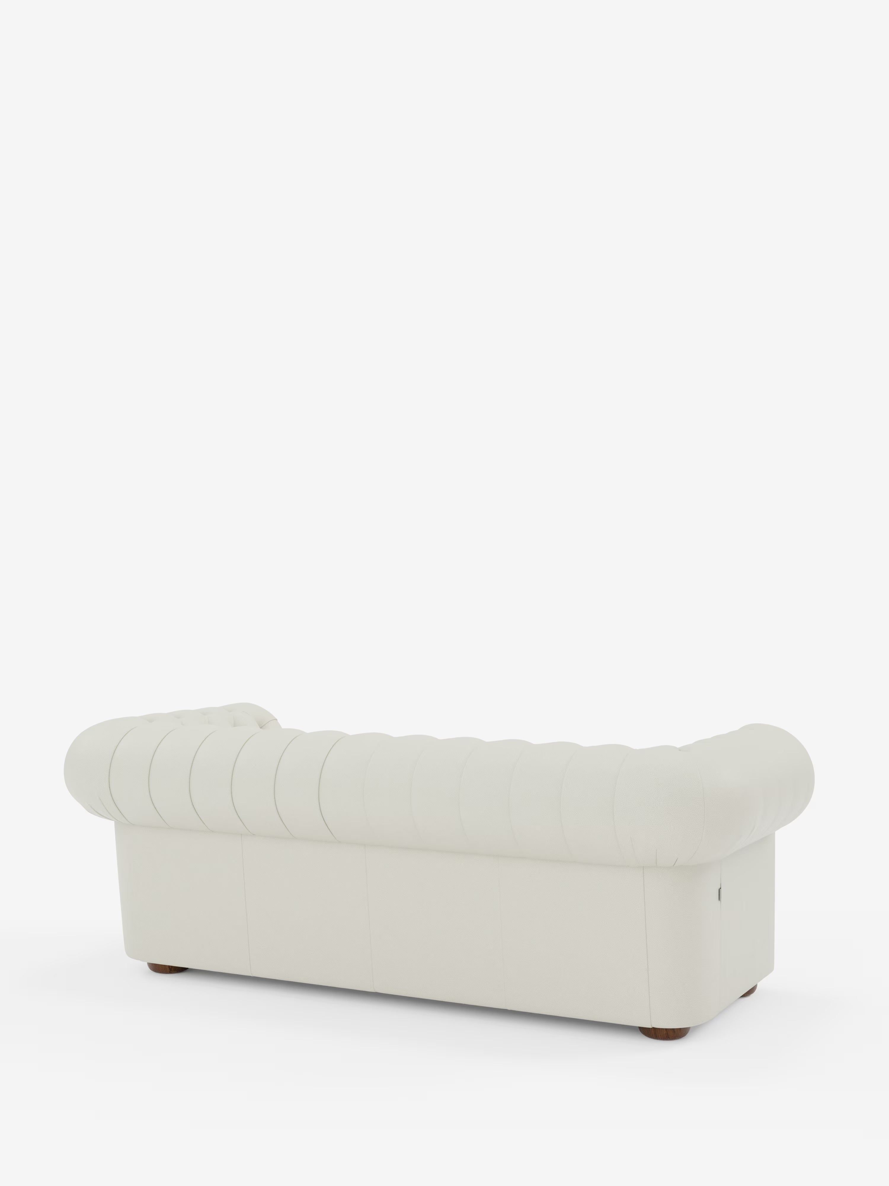 Un divano Chester bianco con capitonné – Divano Chesterfield in vera pelle spessorata a 2 e 3 posti di Divani.Store, caratterizzato da braccioli e schienale arrotondati, è posizionato su uno sfondo bianco a tinta unita. Il divano artigianale è ripreso da dietro e di profilo, con i suoi piedini corti e arrotondati in legno appena visibili sotto.