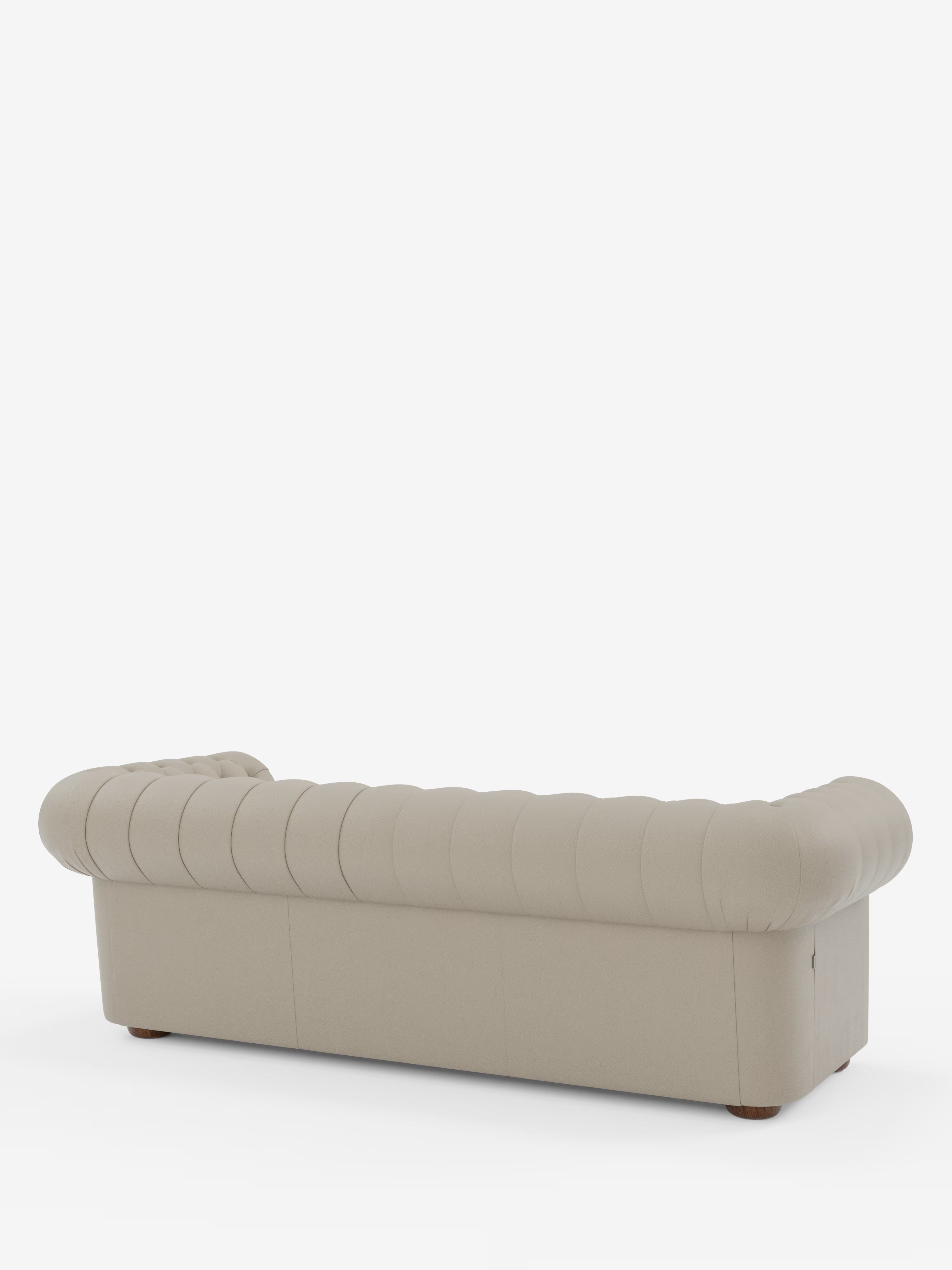 Un divano Chester a 4 posti color beige con tre sedute di Divani.Store, caratterizzato dalla classica lavorazione artigianale Chesterfield Capitonné in vera pelle. Il divano è ripreso di profilo dal retro, mettendo in risalto i braccioli arrotondati e la silhouette sinuosa dal profilo basso su uno sfondo bianco a tinta unita.