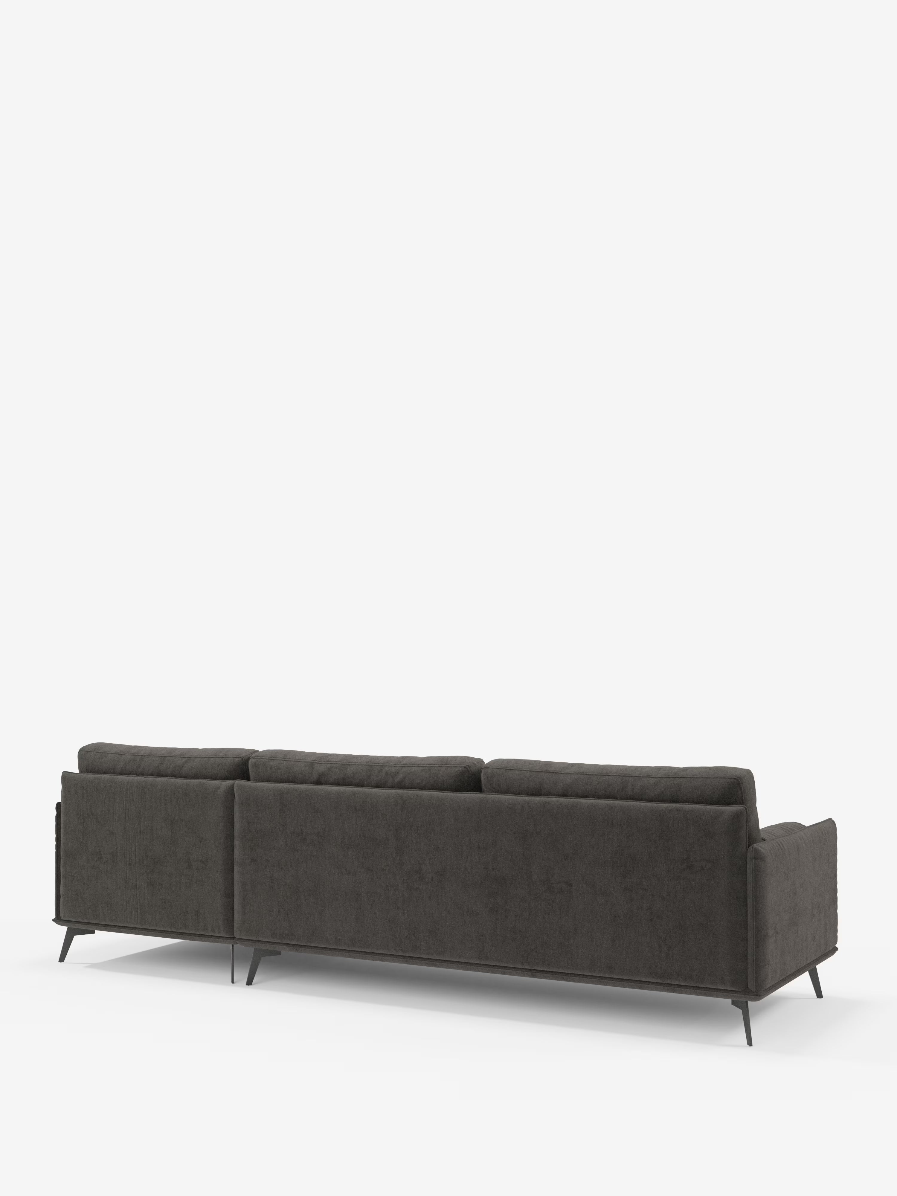 Il divano moderno angolare con penisola Basalto di Divani.Store è presentato nella vista posteriore, rivestito in un moderno tessuto grigio scuro T31, caratterizzato da tre morbidi cuscini dello schienale imbottiti in piuma, sottili gambe angolari in metallo e un design minimalista ed elegante su uno sfondo bianco a tinta unita. Questo elegante divano moderno coniuga perfettamente stile e comfort: perfetto per gli spazi abitativi contemporanei.