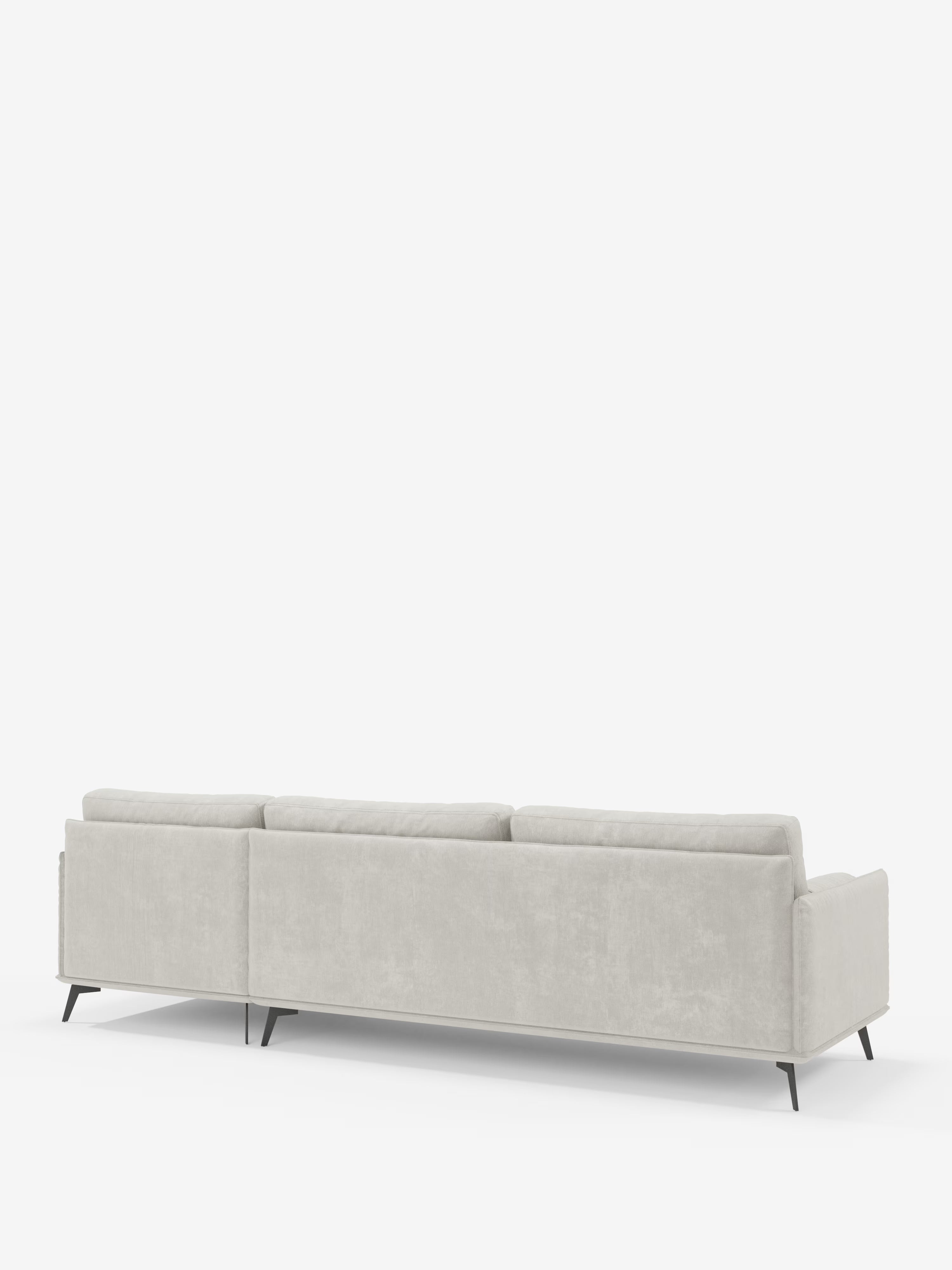 Un divano moderno angolare a tre posti Basalto, di colore grigio chiaro, di Divani.Store, è raffigurato di retro su uno sfondo bianco a tinta unita. Questo divano in tessuto sfoderabile presenta linee pulite, morbidi cuscini in piuma rivestiti in tessuto T31 antimacchia e sottili gambe in metallo nero, per un look contemporaneo e minimalista.