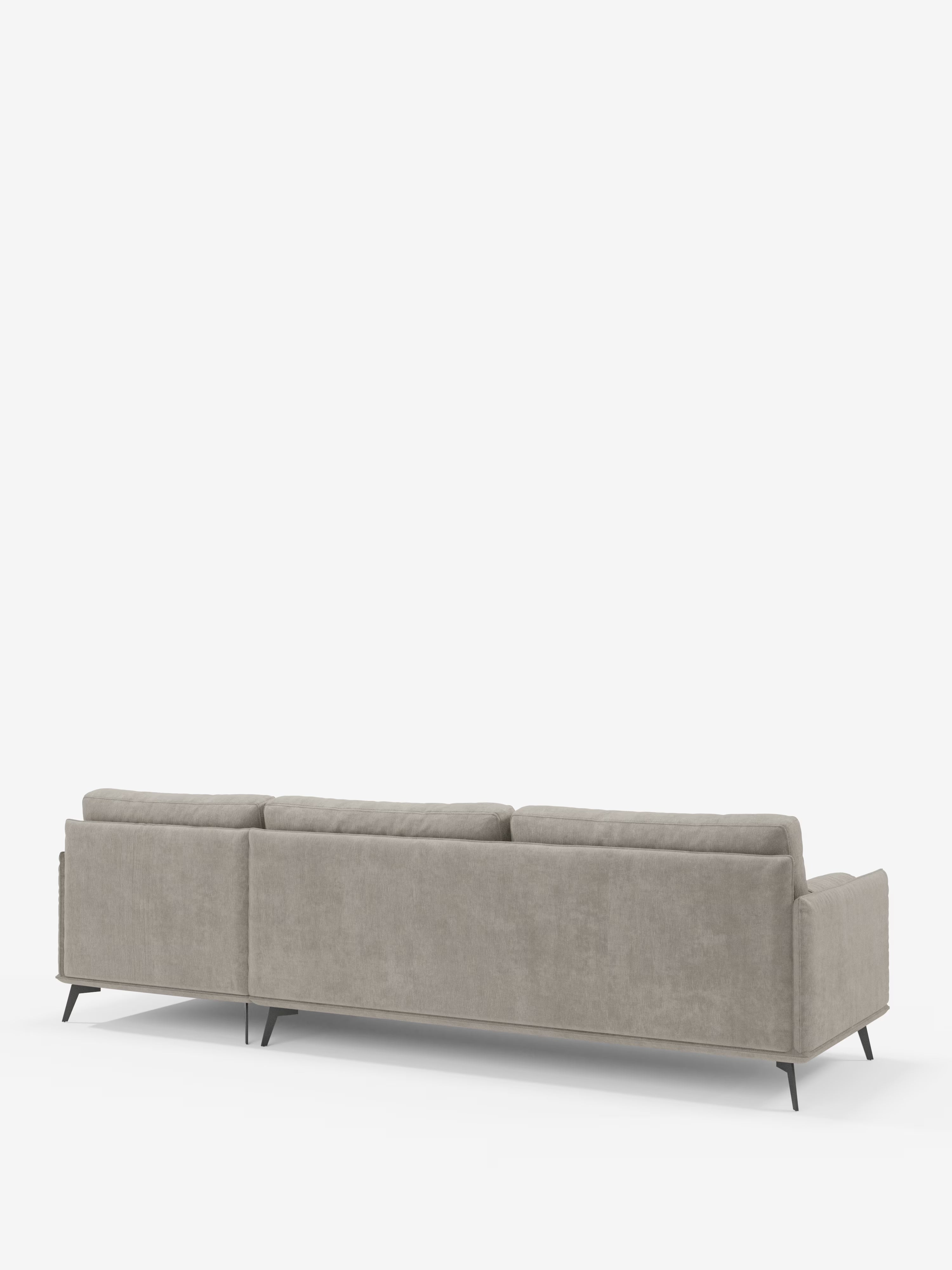 Su uno sfondo bianco spicca il divano angolare moderno "Basalto" di Divani.Store, in grigio chiaro, dotato di cuscini bassi e quadrati imbottiti in piuma e rivestiti in tessuto T31 antimacchia, nonché di un'elegante base in metallo nero. Visto da dietro, mette in risalto le linee pulite e il design minimalista, fondendo con naturalezza l'eleganza italiana contemporanea con un'eccezionale maestria artigianale.