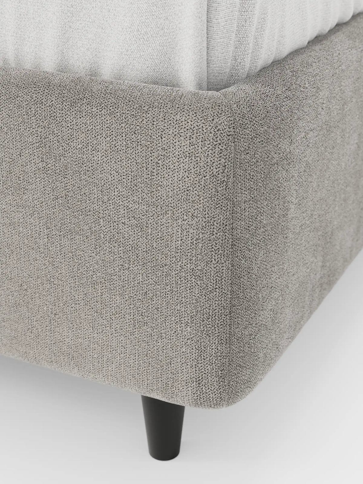 Un primo piano dell'angolo di un letto sommier ERON imbottito, di colore grigio chiaro e con finitura testurizzata, dotato di vano contenitore in tessuto impermeabile certificato Oeko-Tex, che ne mostra la base arrotondata, la gamba cilindrica corta di colore nero e una parte del materasso bianco.