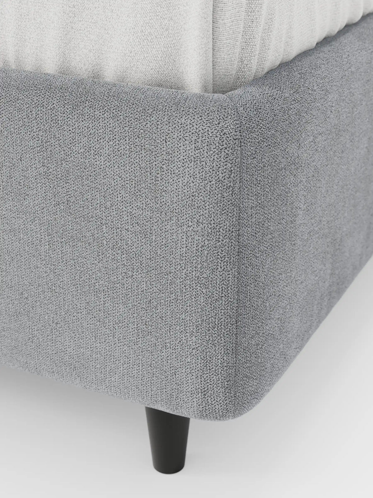 Primo piano di un letto sommier imbottito ERON grigio con vano contenitore in tessuto impermeabile certificato Oeko-Tex, che mostra il tessuto a trama, un materasso bianco e una gamba cilindrica scura su uno sfondo bianco.