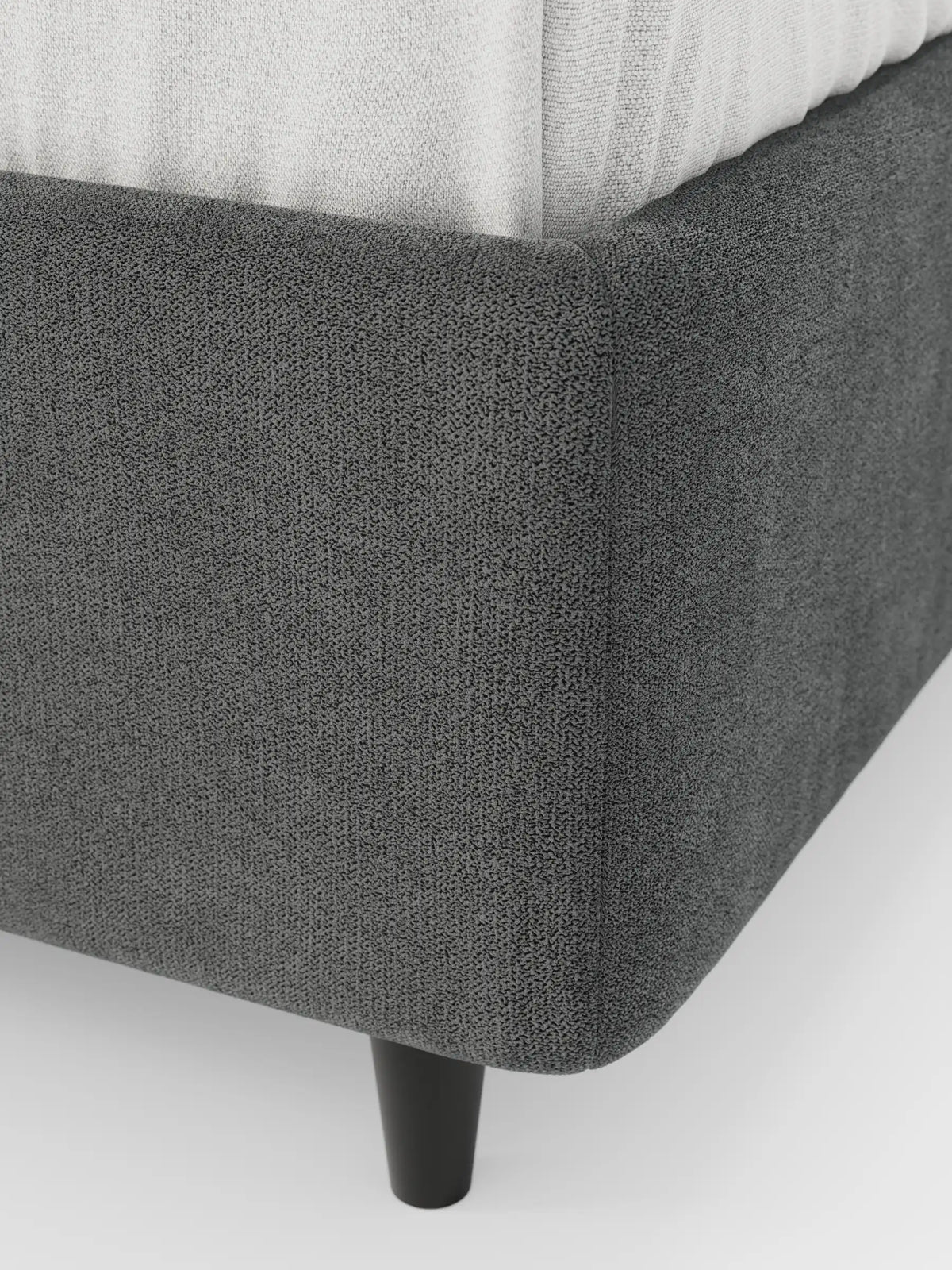 Un primo piano di un letto sommier ERON color grigio antracite, imbottito e dotato di vano contenitore in tessuto impermeabile certificato Oeko-Tex con angoli arrotondati, che mostra la base testurizzata su una gamba nera e una parte di un materasso bianco.