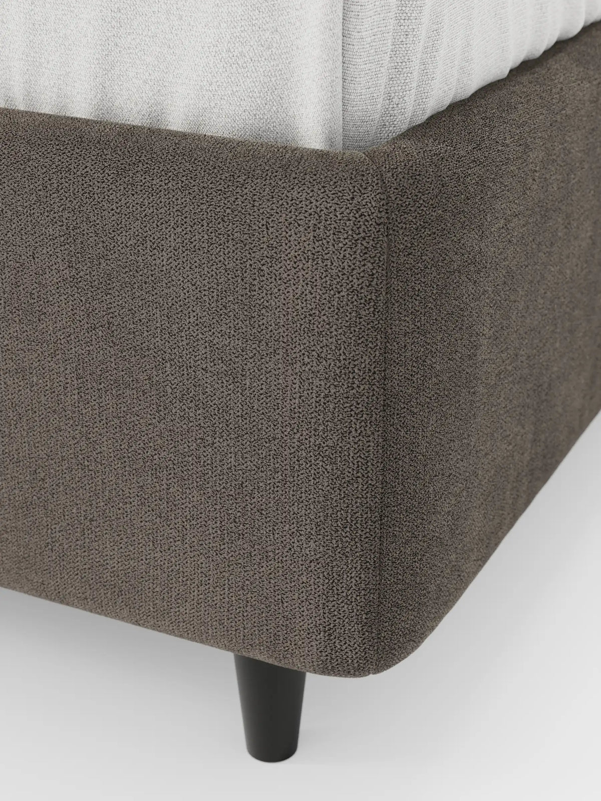 Un primo piano dell'angolo di un letto sommier ERON color marrone scuro, imbottito e dotato di vano contenitore in tessuto impermeabile certificato Oeko-Tex. Il letto poggia su una semplice gamba cilindrica nera, con il materasso bianco visibile sulla parte superiore.