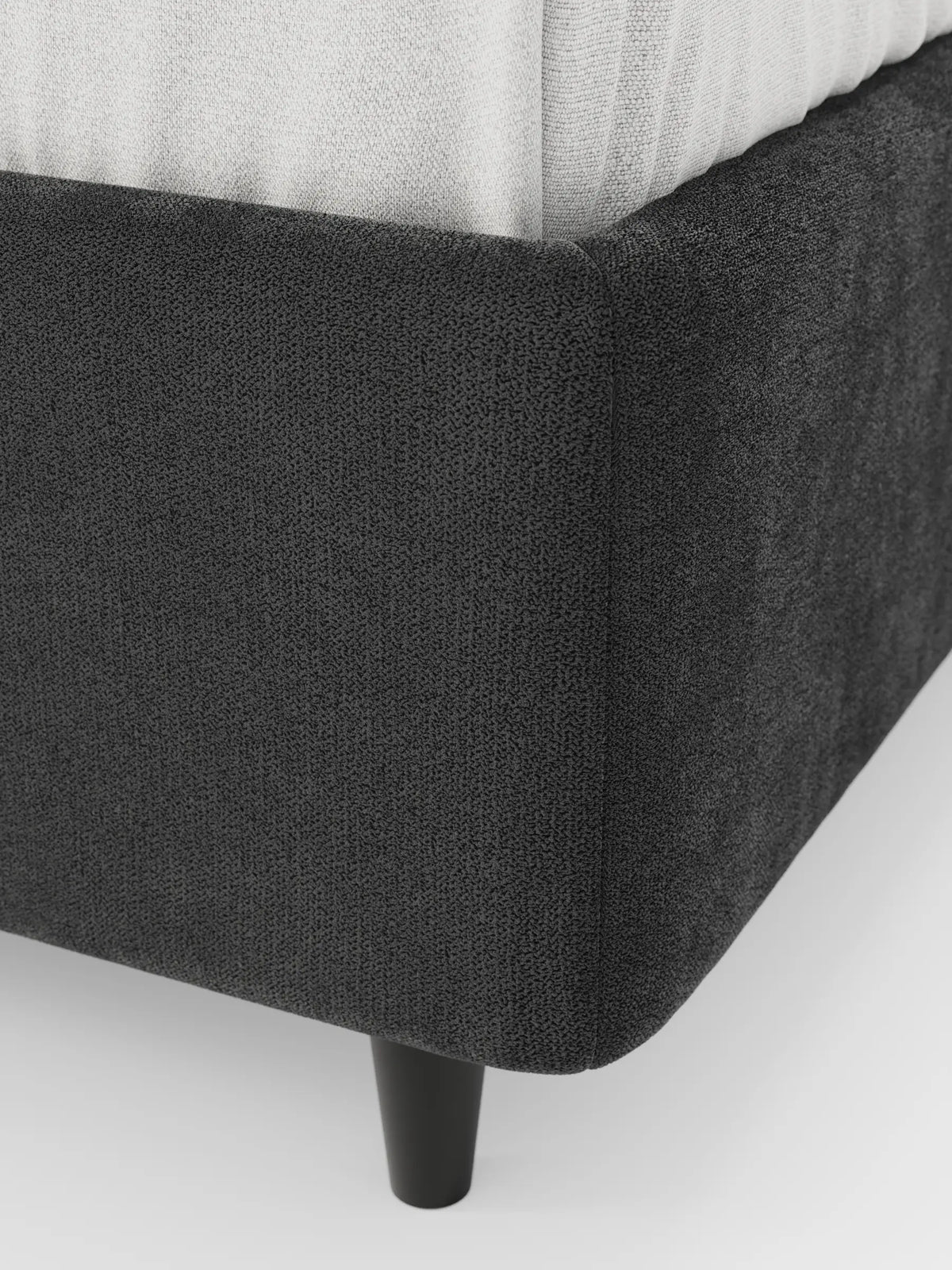 Primo piano del letto sommier imbottito ERON con contenitore, realizzato in tessuto impermeabile certificato Oeko-Tex, caratterizzato da un angolo in tessuto testurizzato grigio scuro, una gamba cilindrica nera e un materasso grigio chiaro su sfondo bianco.