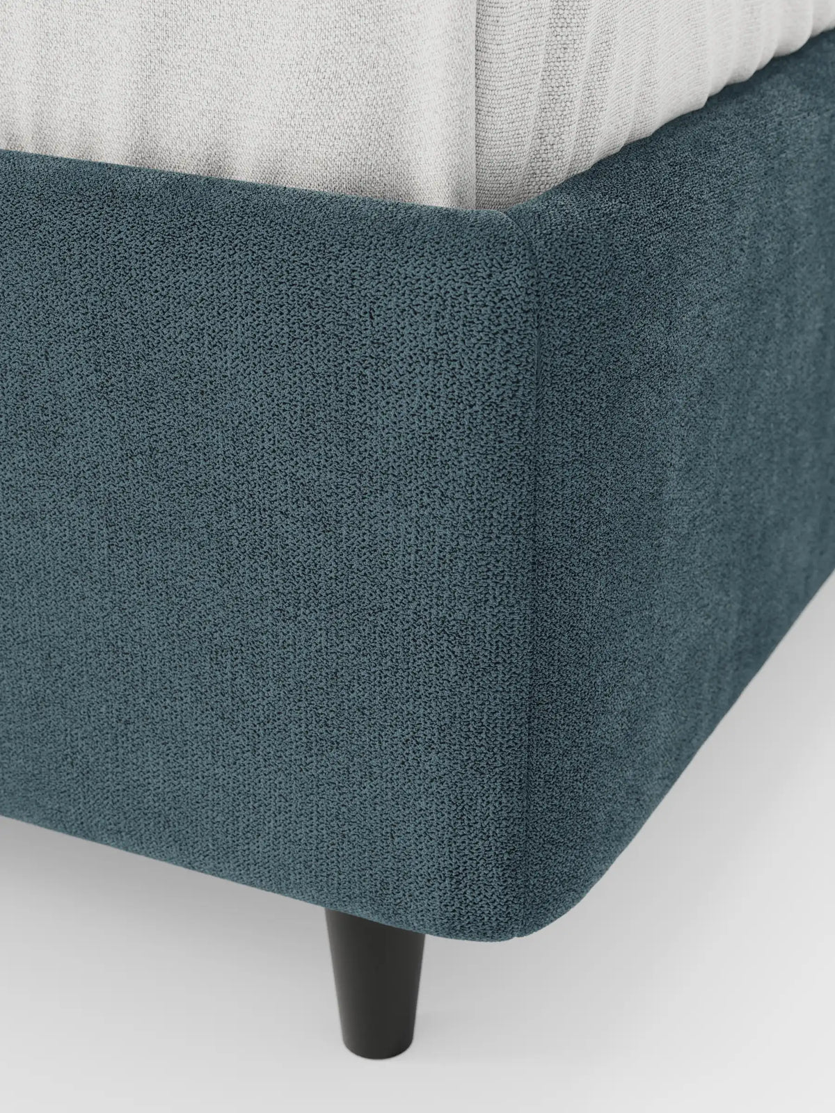 Un primo piano dell'angolo di un letto sommier imbottito ERON con vano contenitore in tessuto impermeabile certificato Oeko-Tex color verde acqua scuro, con un materasso bianco e una gamba cilindrica corta e scura su sfondo bianco.