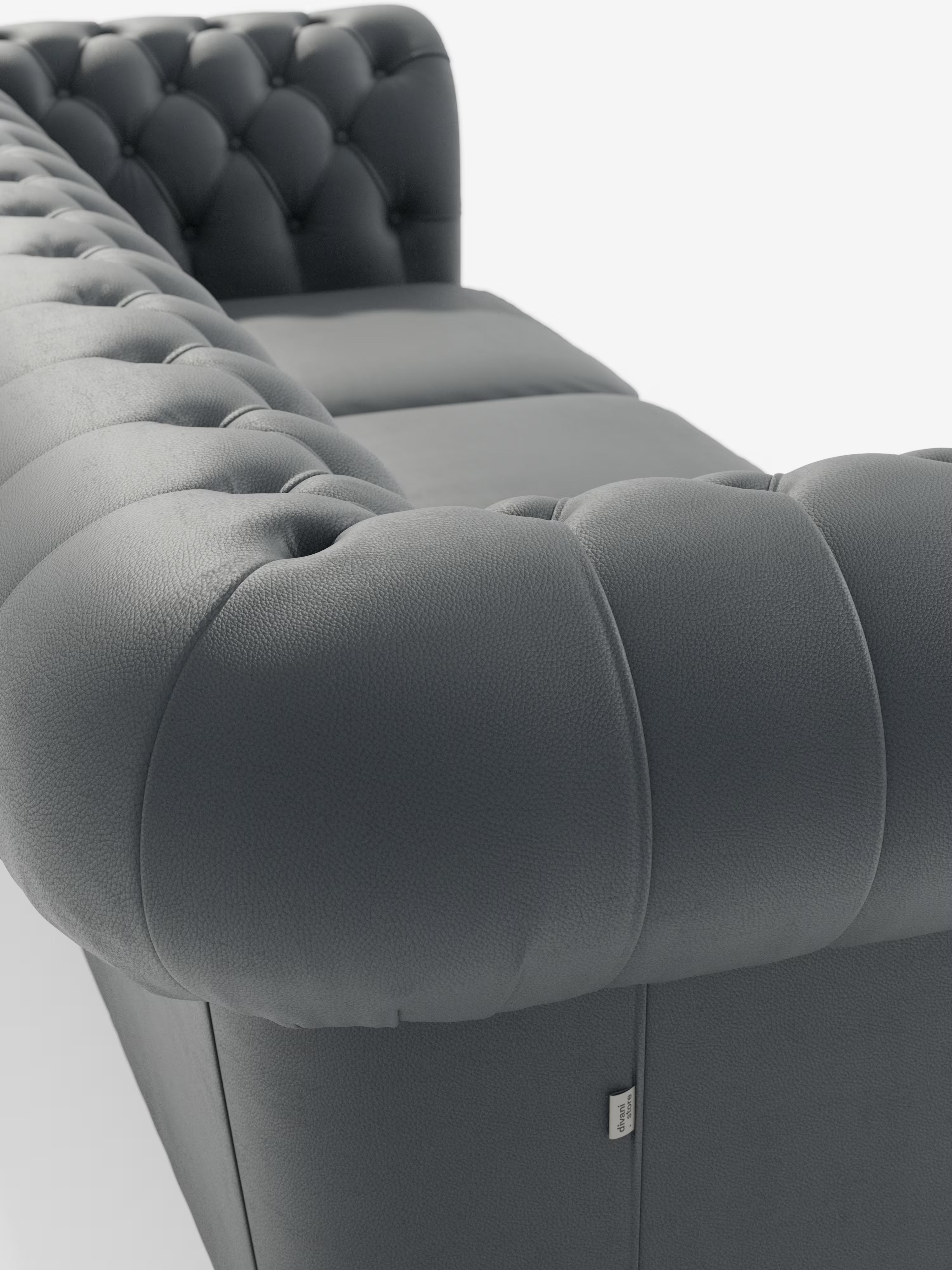 Primo piano del Divani.Store Chester 4 Posti 3 sedute in Vera Pelle – Chesterfield Capitonné Artigianale, un divano in pelle capitonné grigio scuro con braccioli arrotondati, che richiama lo stile intramontabile di un classico divano Chesterfield. La vera pelle finemente lavorata e le cuciture meticolose ne sottolineano la lavorazione artigianale, mentre sul lato anteriore è applicata una piccola etichetta in tessuto. Il divano è posizionato su uno sfondo bianco a tinta unita.