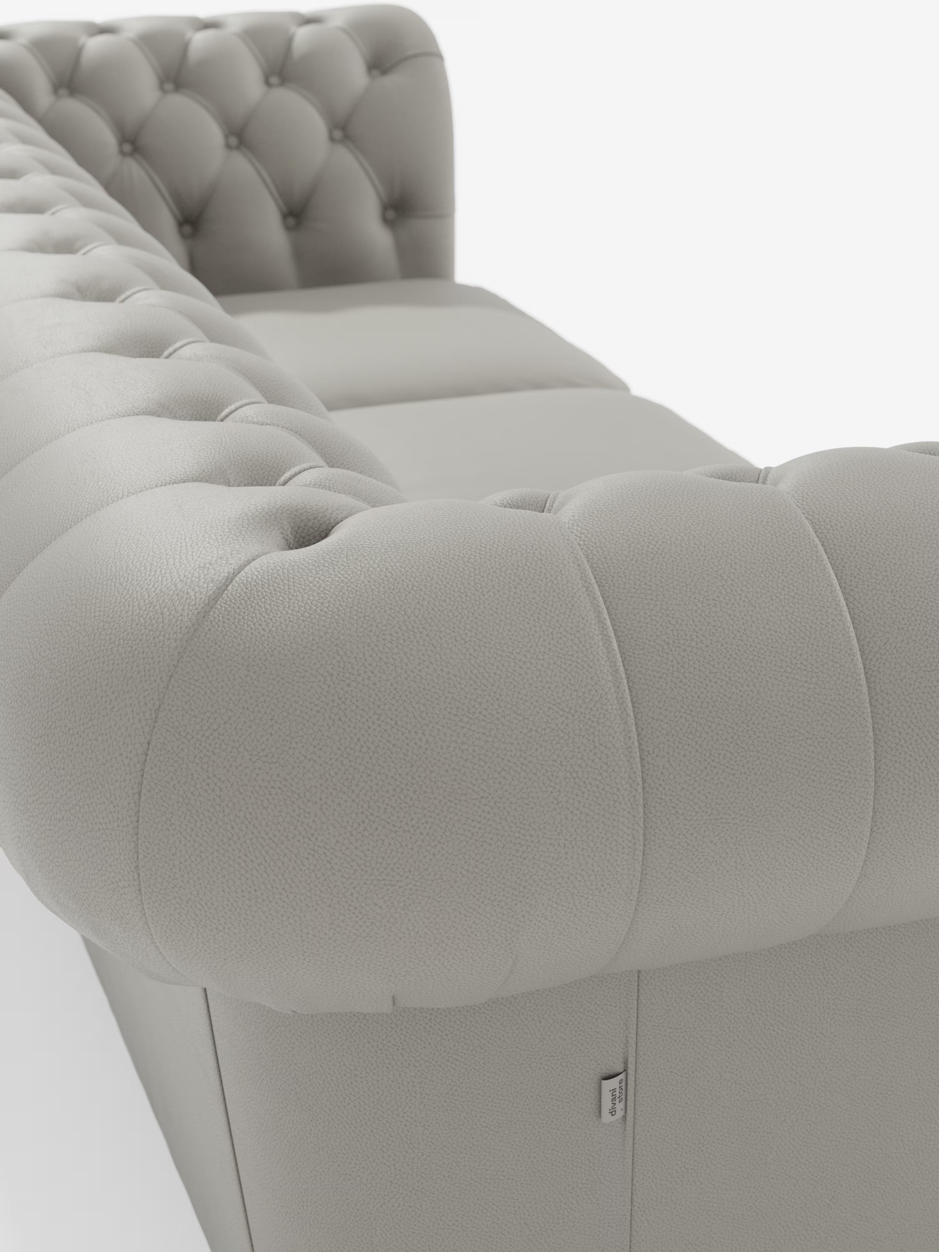 Primo piano del Chester 4 Posti 3 sedute in Vera Pelle – Chesterfield Capitonné Artigianale di Divani.Store, un divano in pelle grigio chiaro con finitura capitonné e profondi bottoni decorativi sui braccioli e sullo schienale, ripreso da un’angolazione laterale. Il rivestimento, realizzato con maestria, presenta una finitura testurizzata; nella parte inferiore è presente una piccola etichetta in tessuto bianco con scritta nera.