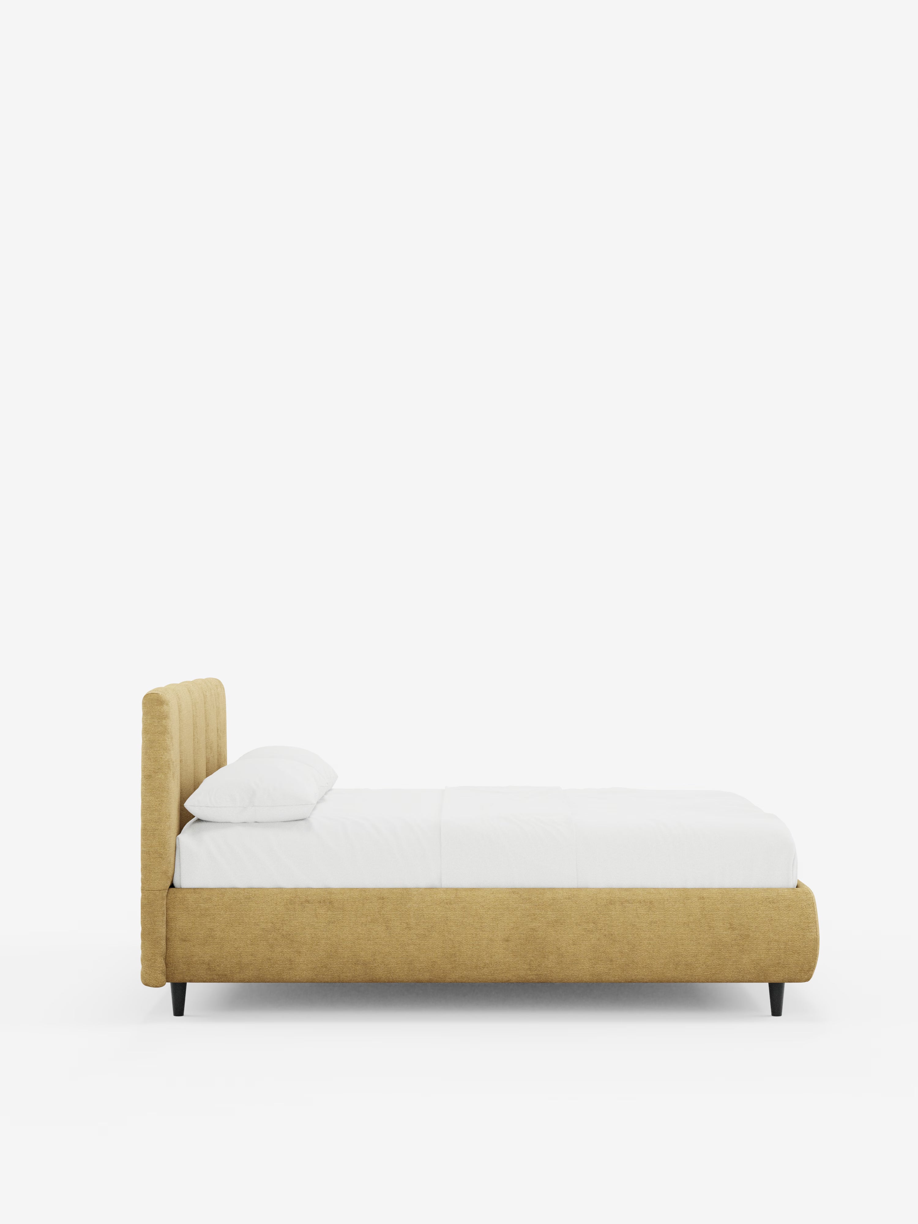 Vista laterale del letto imbottito ARKAM con contenitore e testiera a lavorazione verticale di Letti.store, caratterizzato da una struttura in tessuto giallo senape certificato Oeko-Tex®, una testiera bassa con cuciture verticali in stile shabby chic, biancheria da letto bianca con un unico cuscino bianco; il letto poggia su gambe corte di colore scuro su uno sfondo bianco a tinta unita.