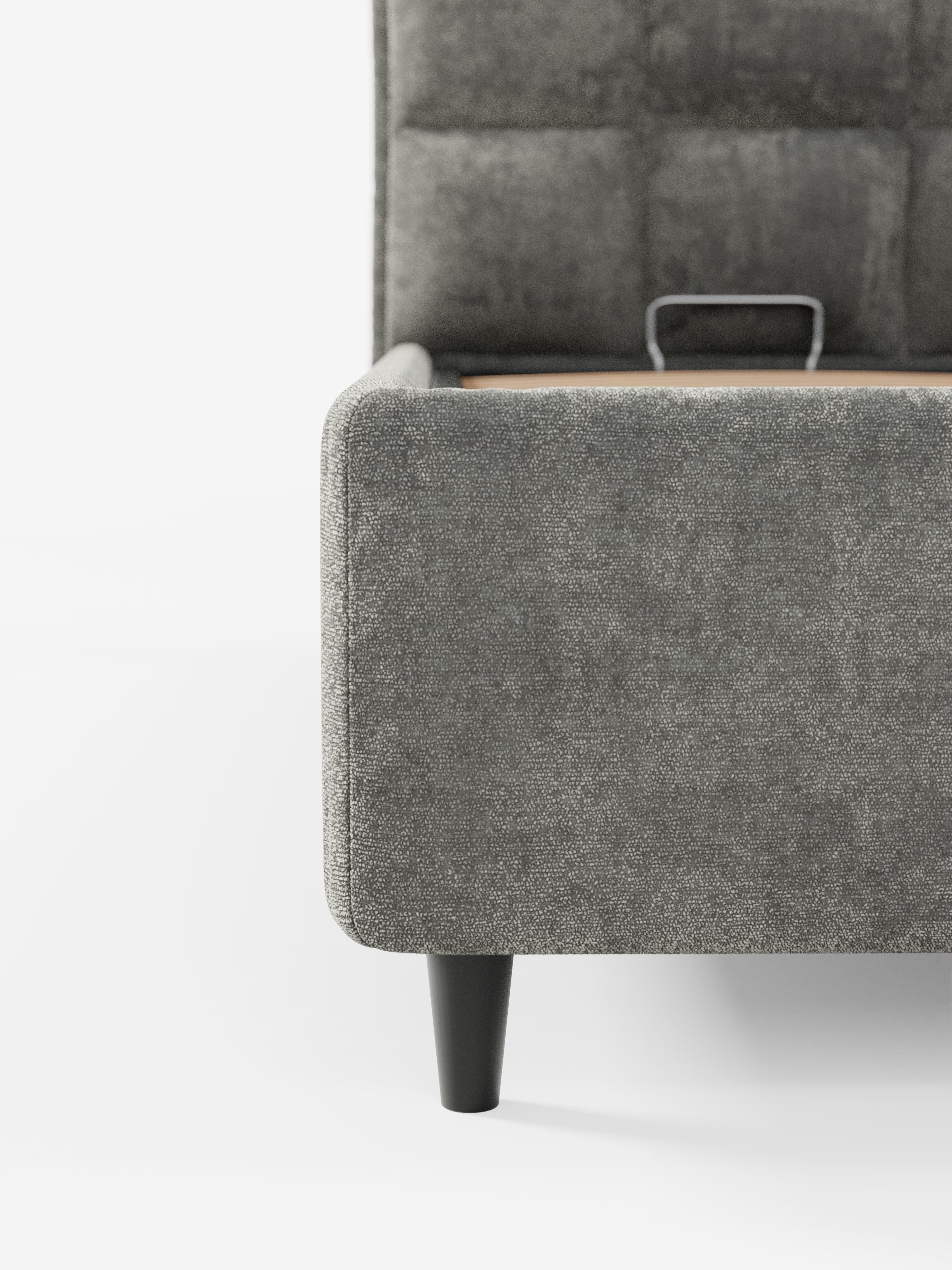 Primo piano di un letto imbottito AURO di Letti.store, caratterizzato da un angolo della struttura del letto rivestito in ciniglia grigia certificata Oeko-Tex® con tessuto testurizzato e da una gamba cilindrica in legno nero. È visibile parte della testiera alta e capitonné. Una piccola maniglia in metallo vicino al centro indica la presenza di un pratico vano contenitore per una maggiore praticità di stoccaggio. Lo sfondo è bianco a tinta unita.