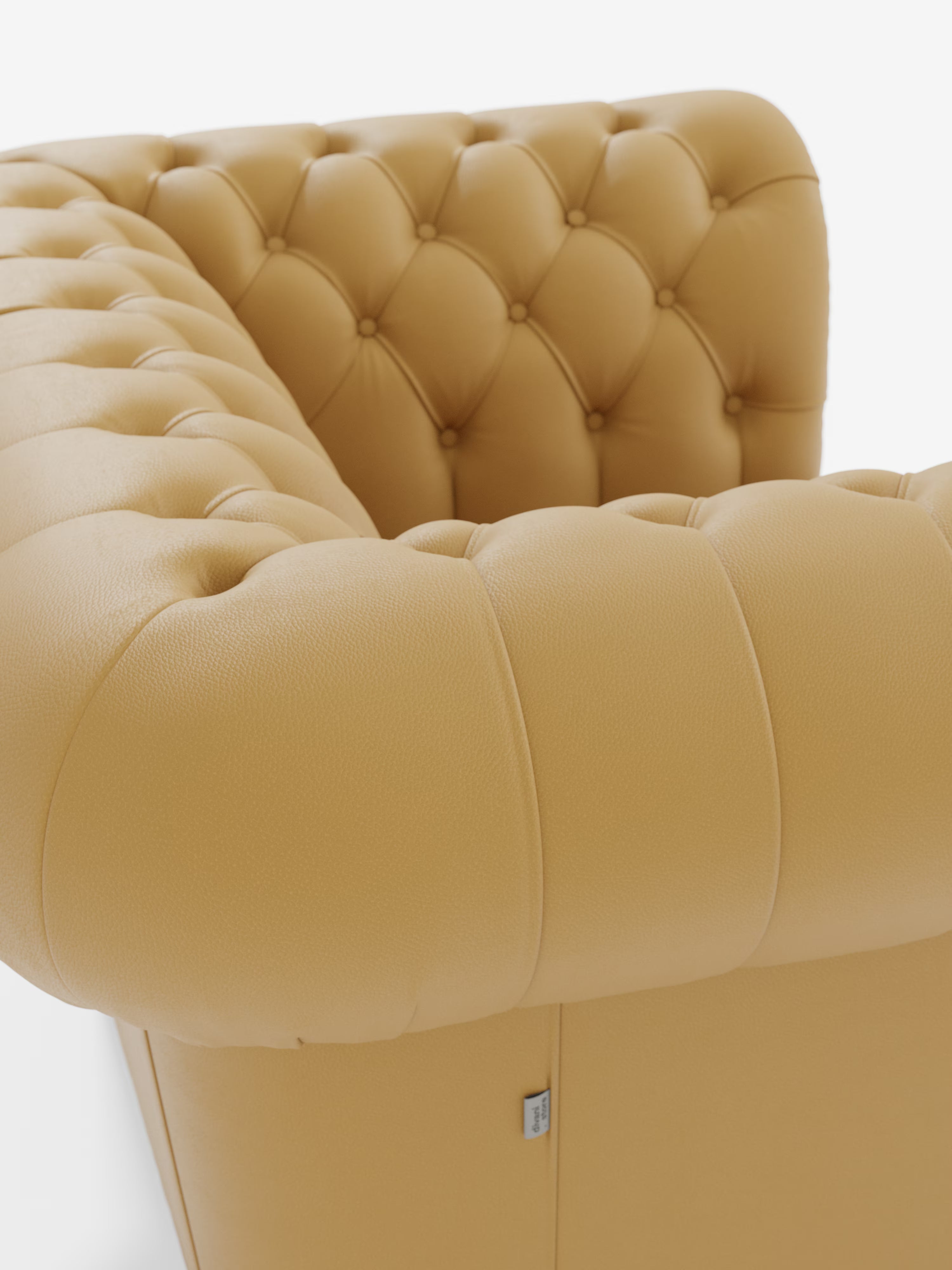 Primo piano del bracciolo e dello schienale della poltrona Chester di Divani.Store, realizzata in vera pelle spessorata con capitonné artigianale, che mette in risalto il rivestimento in pelle color senape con bottoni a vista e una superficie dalla trama ricca che evoca un’eleganza raffinata. Vicino alla base è visibile una piccola etichetta in tessuto recante il marchio Divani.Store, che ne esalta il fascino lussuoso. Lo sfondo è bianco a tinta unita.