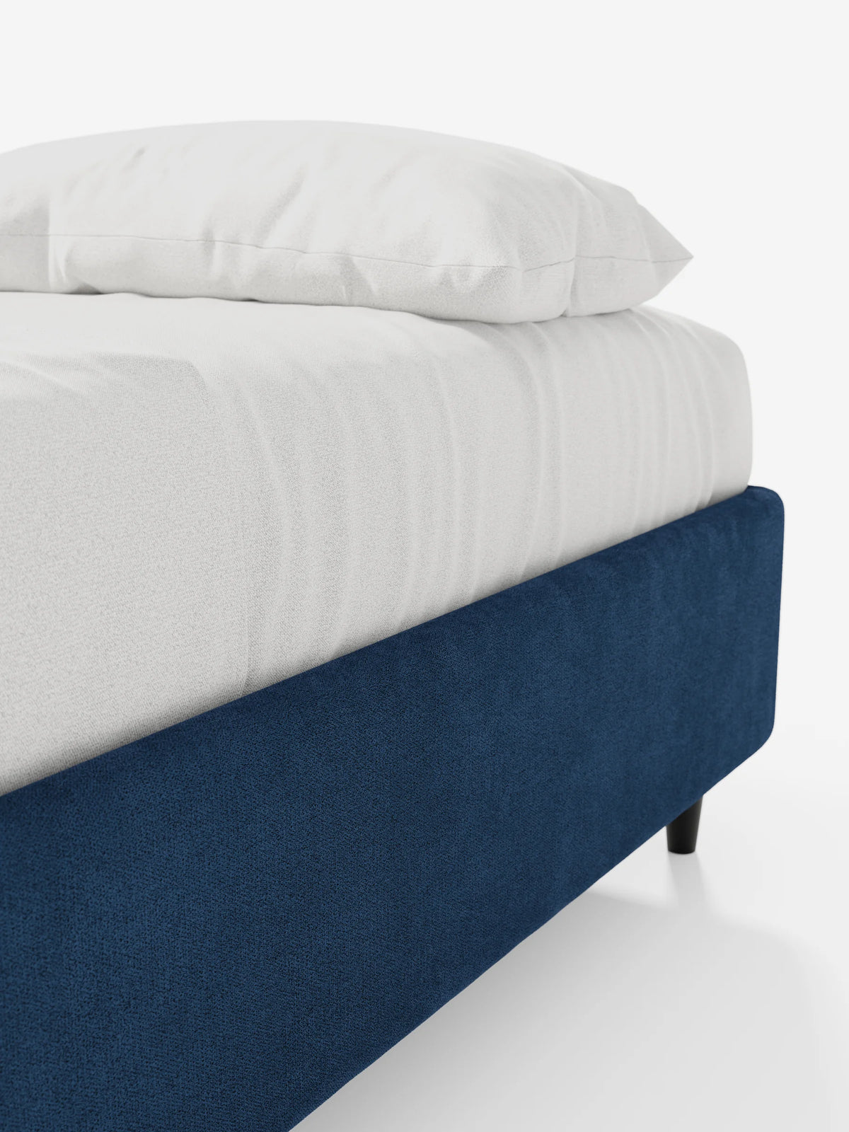Un primo piano del letto sommier imbottito ERON con contenitore, rivestito in tessuto impermeabile certificato Oeko-Tex di colore blu scuro, con un lenzuolo e un cuscino bianchi immacolati appoggiati su gambe basse e affusolate di colore scuro, su uno sfondo bianco a tinta unita.