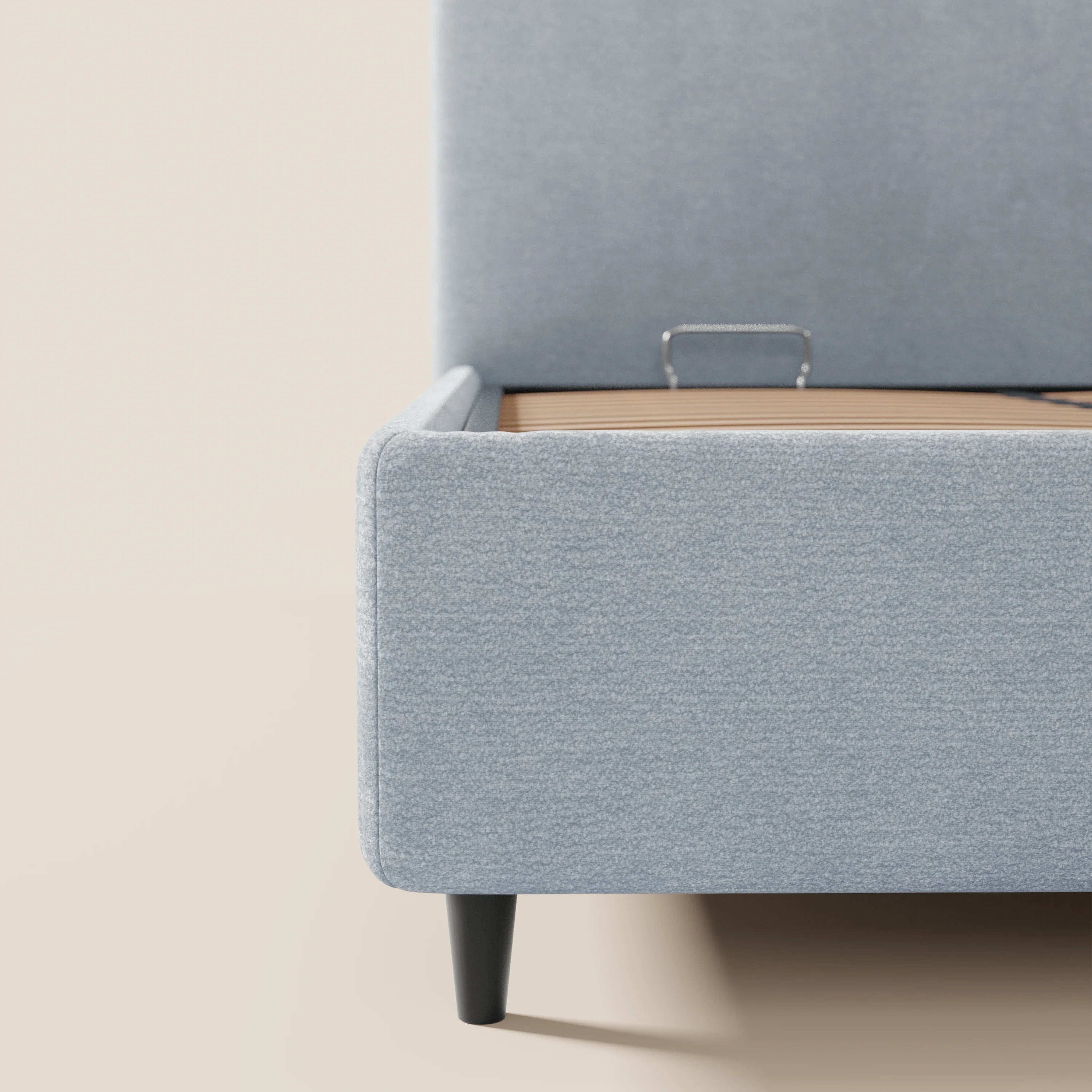 Primo piano del letto KAELOS minimal imbottito di Letti.store in ciniglia azzurra, dotato di base contenitiva con maniglia in metallo, gambe alte nere e testiera arrotondata. La foto mette in risalto l'angolo in basso a destra su uno sfondo bianco sporco.