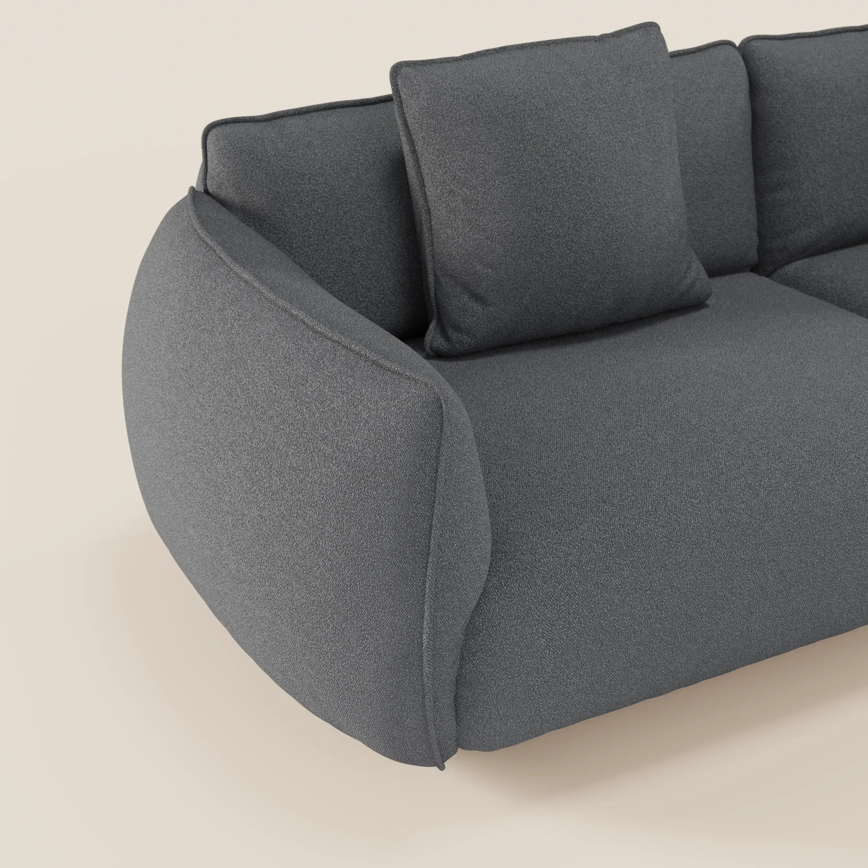 Primo piano del divano Nimbus 3 posti modulare e componibile di Divani.Store in tessuto smacchiabile effetto cotone grigio scuro, con braccioli arrotondati e due cuscini quadrati, su sfondo beige chiaro. Solo una parte è visibile.