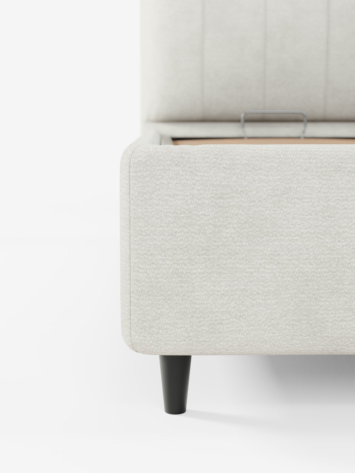 Primo piano di un letto imbottito ONYX con vano contenitore e testiera verticale in tessuto certificato Oeko-Tex® di Letti.store, che mostra l’angolo della struttura del letto imbottito grigio chiaro con il coperchio del vano contenitore parzialmente aperto, lasciando intravedere le finiture in legno e una maniglia in metallo. È visibile una singola gamba nera affusolata, in risalto su uno sfondo bianco liscio.