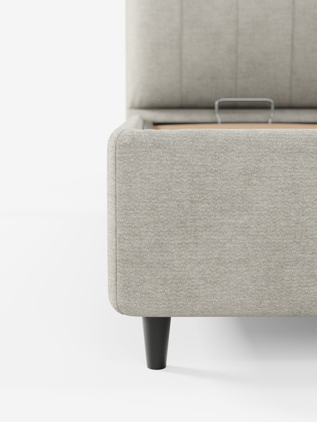 Primo piano del letto imbottito ONYX con vano contenitore e testiera verticale in tessuto certificato Oeko-Tex® di Letti.store, caratterizzato da un’angolo della struttura imbottita in grigio chiaro con piedini corti in legno nero e una discreta maniglia in metallo sulla parte superiore, che ne rivela il vano contenitore nascosto. Lo sfondo bianco e minimalista mette in risalto le linee pulite e l’eleganza contemporanea di questo letto moderno con testiera verticale imbottita.