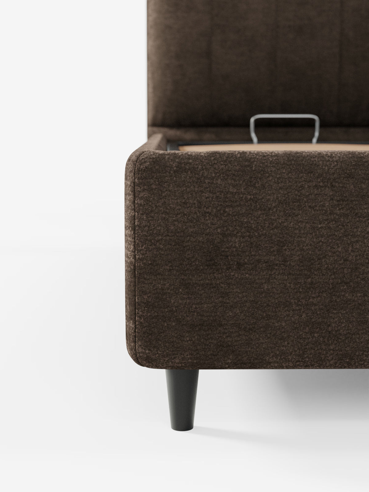 Un primo piano del letto imbottito ONYX con vano contenitore e testiera verticale in tessuto certificato Oeko-Tex® di Letti.store mette in risalto un angolo della struttura del letto imbottito marrone con una gamba affusolata nera. Il coperchio del letto è leggermente sollevato, rivelando all’interno una maniglia in metallo che consente di accedere al vano contenitore integrato. Lo sfondo è bianco a tinta unita.
