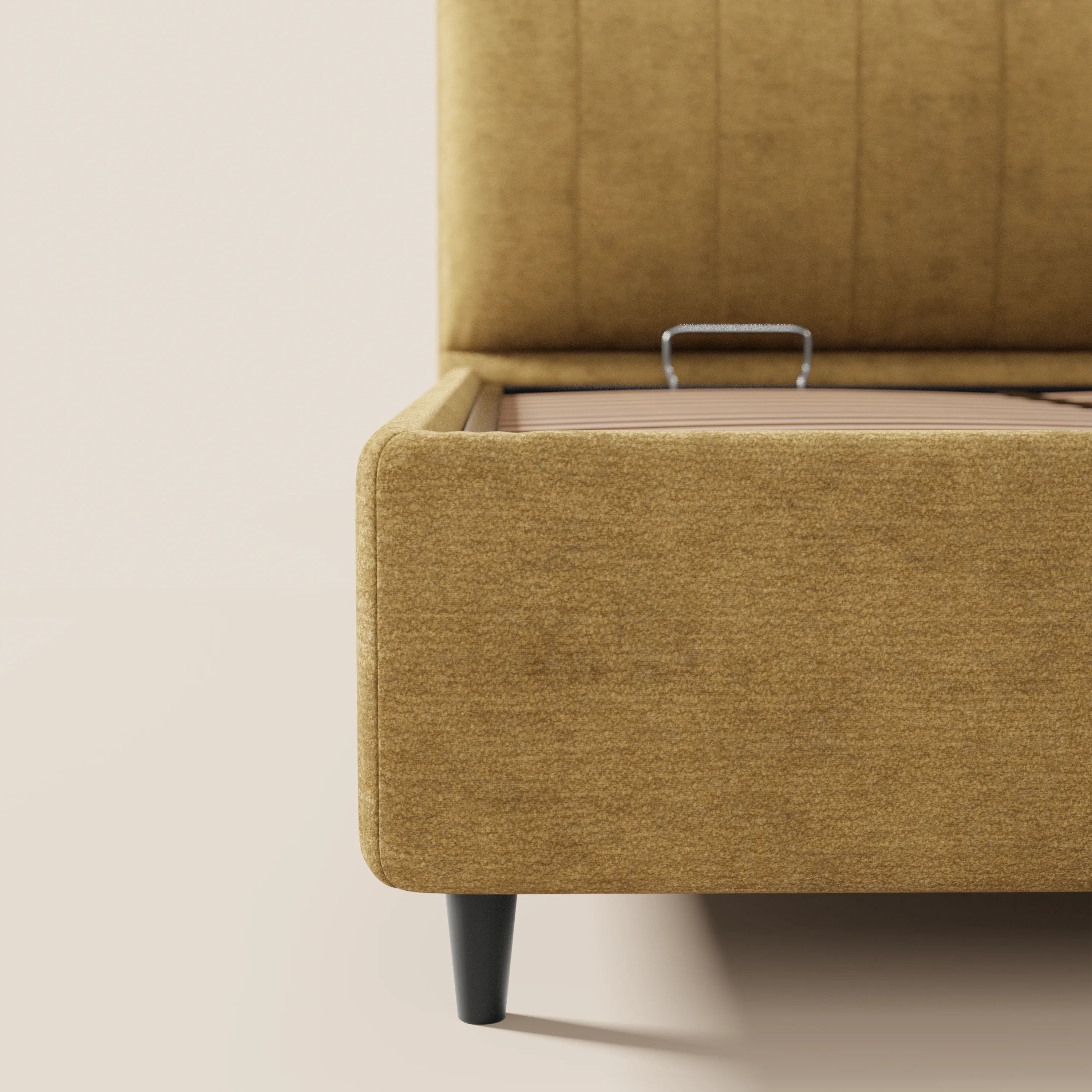 Primo piano del letto imbottito ONYX con contenitore di Letti.store, realizzato in tessuto Oeko-Tex®: giallo senape, gambe affusolate nere, materasso sollevato per mostrare il vano contenitore con maniglia argentata. Lo sfondo è a tinta unita e di colore chiaro.