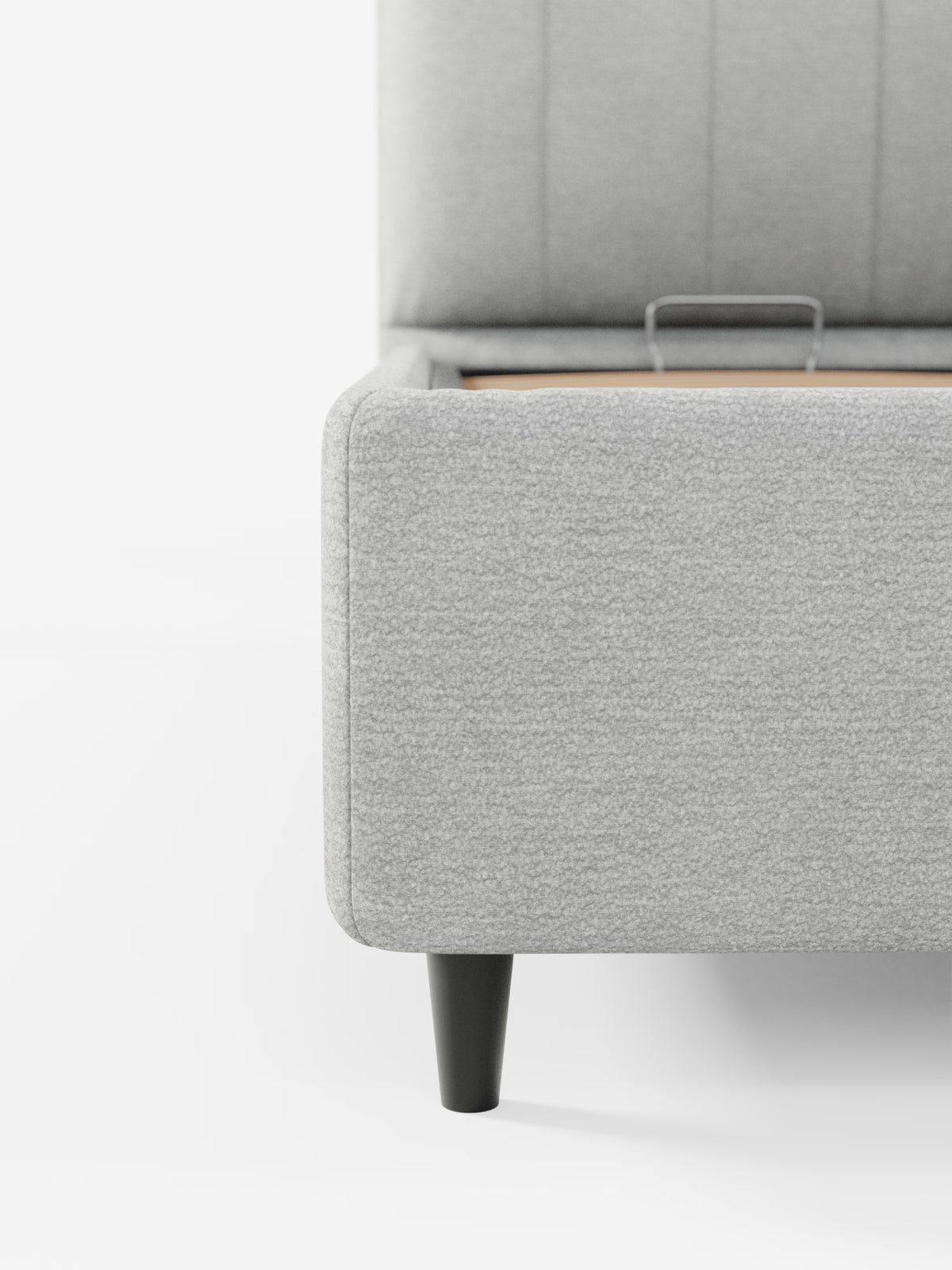 Primo piano del letto imbottito ONYX con contenitore e testiera verticale in tessuto certificato Oeko-Tex® di Letti.Il negozio presenta un letto angolare imbottito grigio chiaro con tessuto dalla trama morbida, una gamba cilindrica nera, cuciture verticali sulla testiera imbottita e un accenno dello spazio interno in legno. Lo sfondo e il pavimento bianchi creano un'estetica pulita e minimalista.