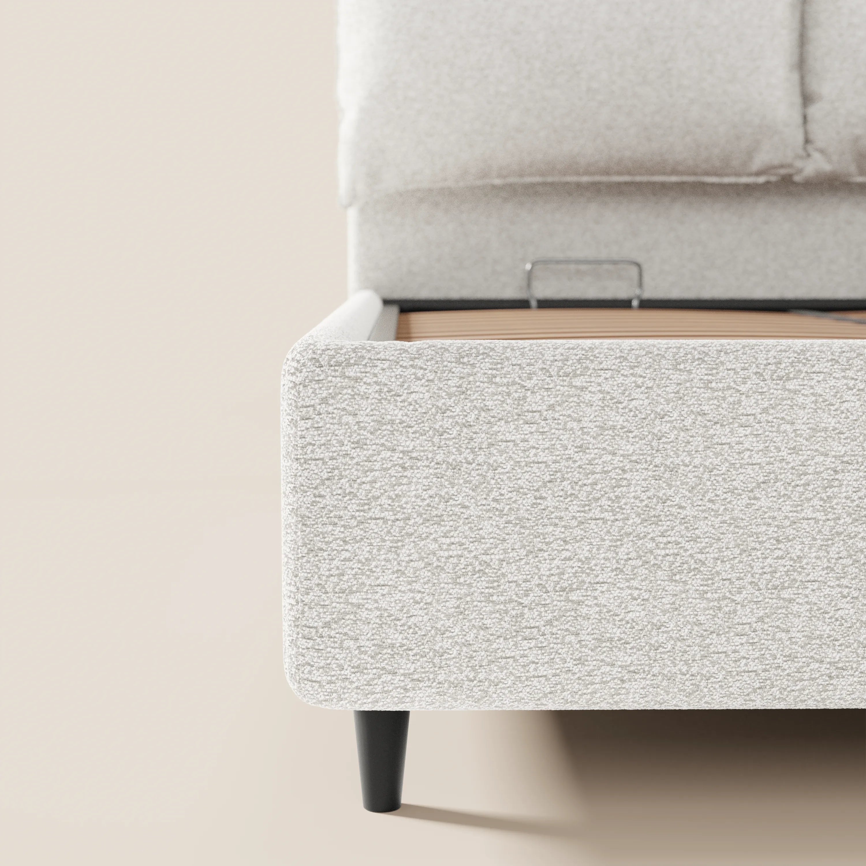 Un primo piano del letto imbottito ZAYRON con contenitore e morbidi cuscini regolabili di Letti.store, realizzato in tessuto grigio chiaro testurizzato certificato Oeko-Tex®, con gambe angolate nere e una testiera alta imbottita su uno sfondo beige chiaro.