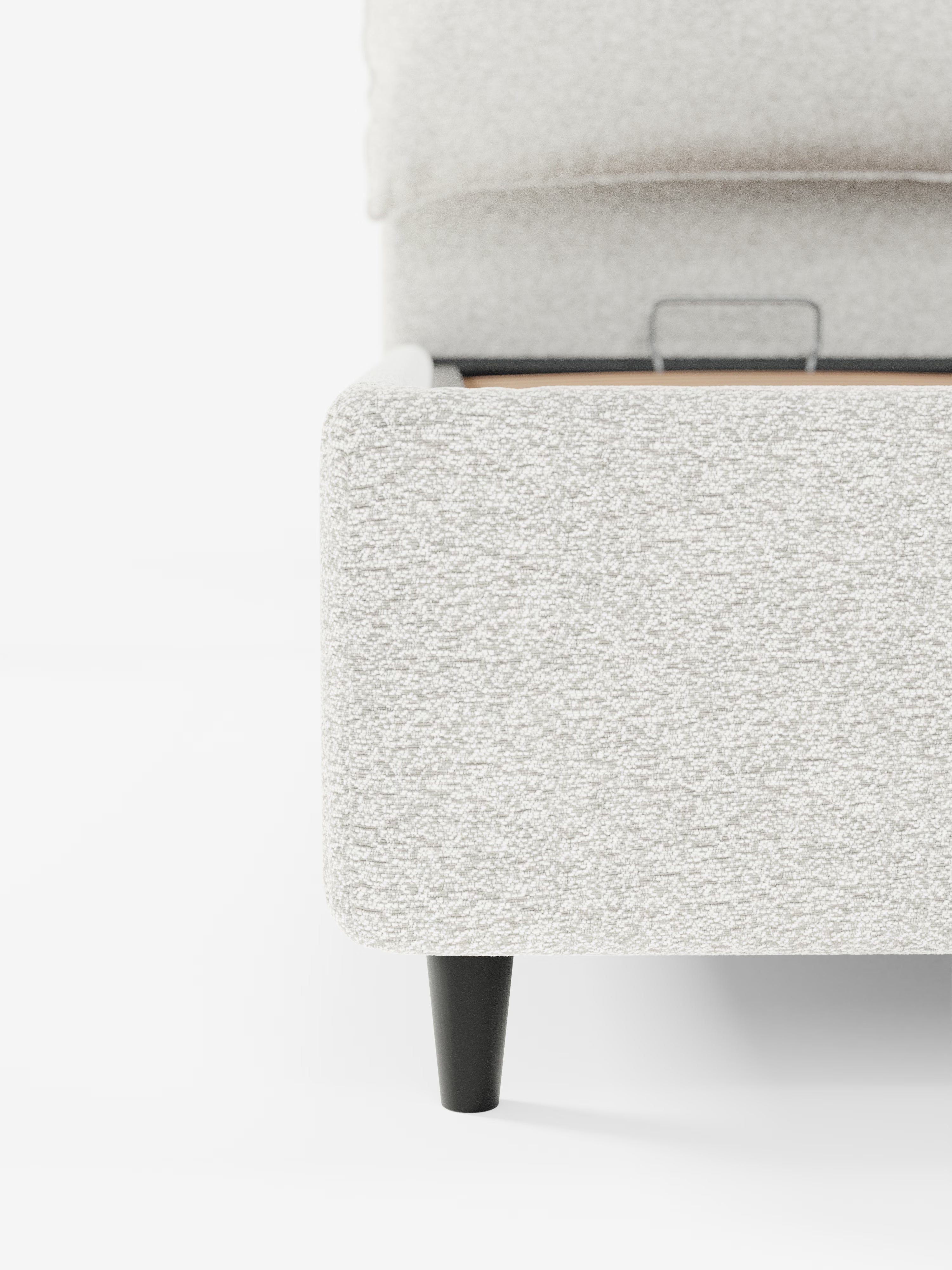 Primo piano di un letto imbottito ZAYRON, dalla trama ruvida e di colore grigio chiaro, con vano contenitore e morbidi cuscini regolabili, realizzato da Letti.store, caratterizzato da un moderno tessuto certificato Oeko-Tex®. L'angolo mostra gambe affusolate nere e una staffa di sostegno in metallo a vista all'interno del telaio. Il tutto su uno sfondo bianco a tinta unita che mette in risalto il tessuto del letto e i dettagli costruttivi. È visibile solo una parte del letto.