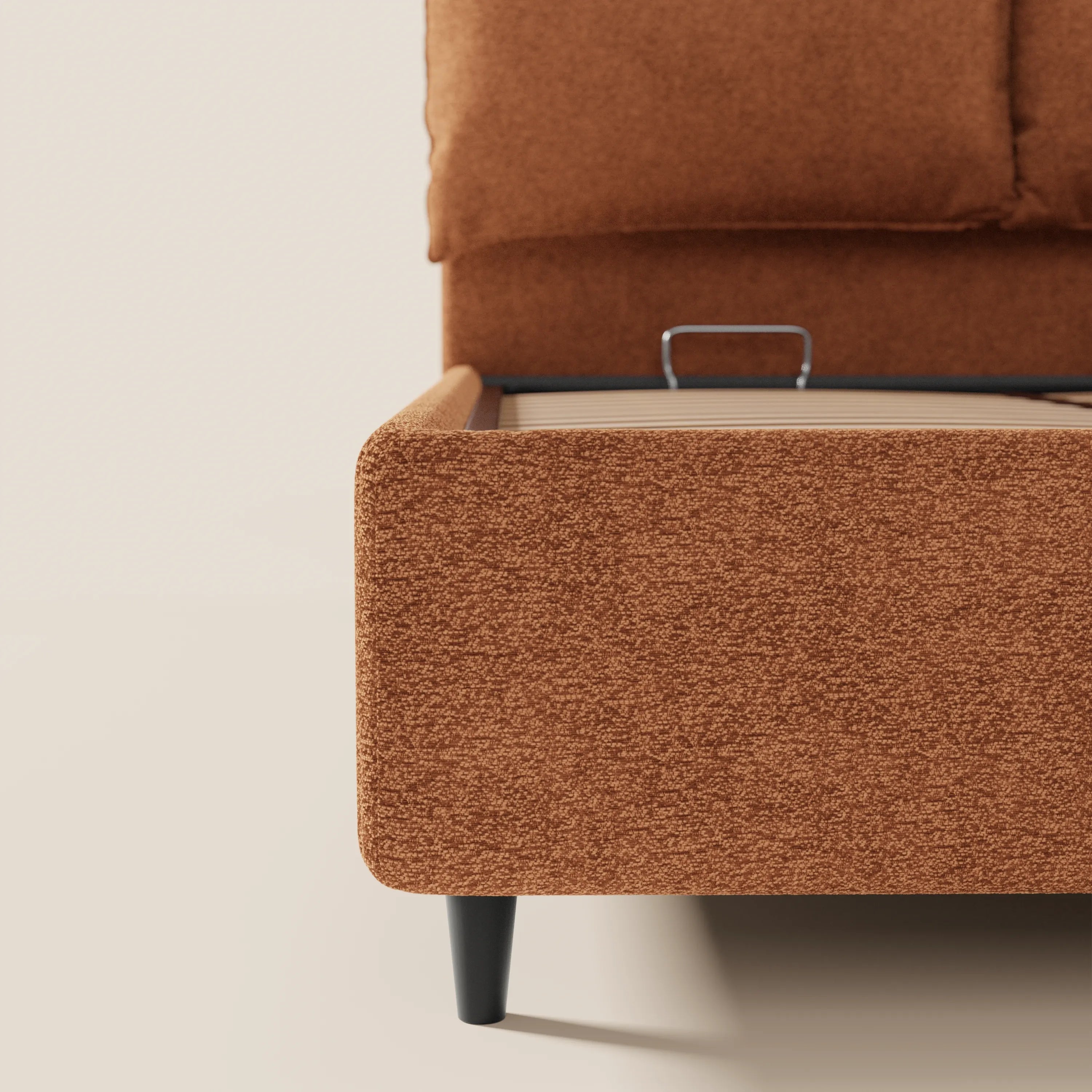 Un primo piano del letto imbottito con contenitore ZAYRON di Letti.store, caratterizzato da un tessuto color ruggine certificato Oeko-Tex®, gambe corte e angolate di colore nero, morbidi bordi arrotondati, una maniglia in metallo sulla base e cuscini regolabili, il tutto su sfondo di pareti e pavimento color bianco sporco.