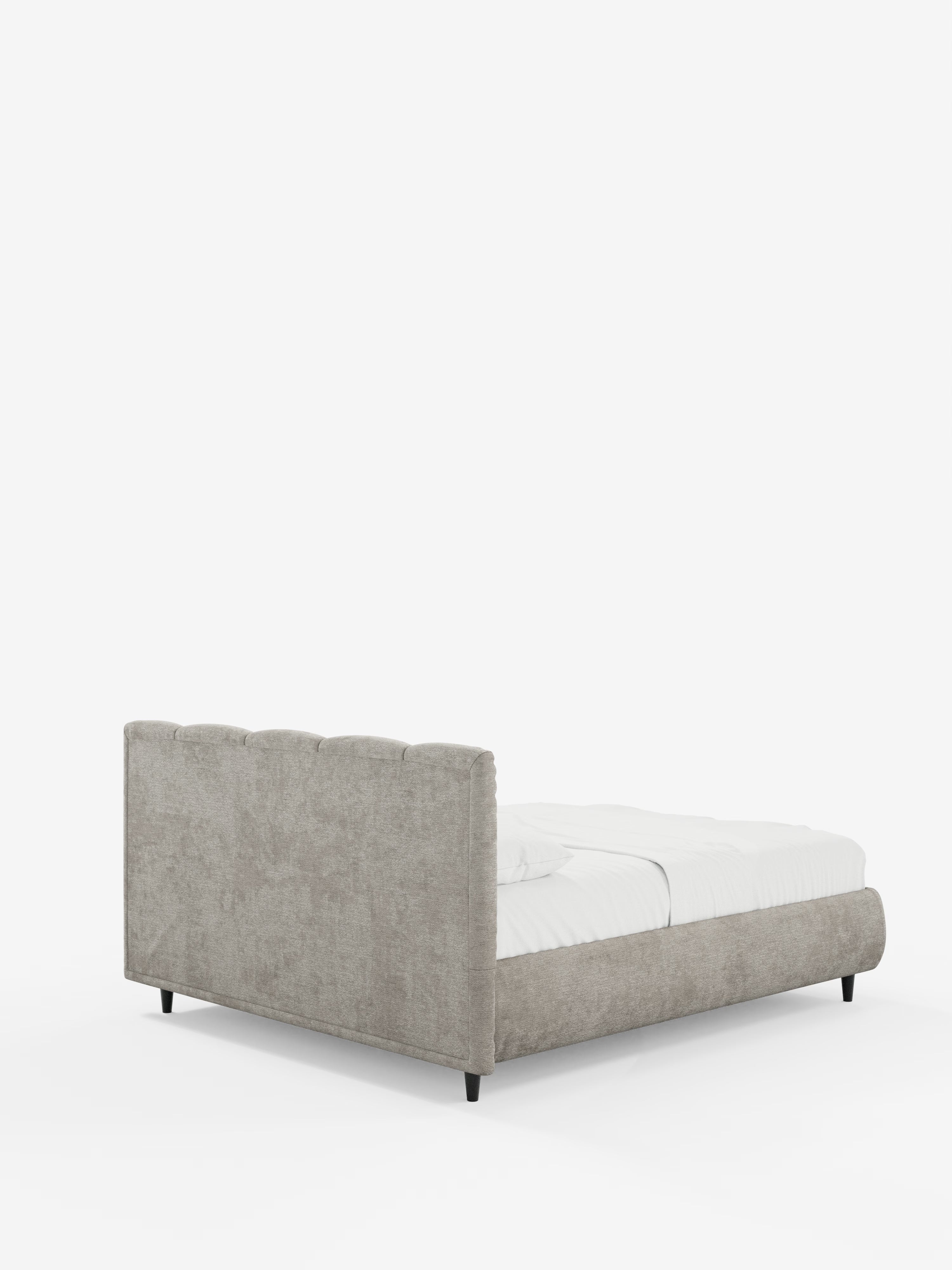 Il letto ARKAM, un modello basso e moderno di Letti.store, imbottito, con vano contenitore e testiera a cuciture verticali, rivestito in tessuto beige chiaro certificato Oeko-Tex®, con gambe nere e biancheria da letto bianca, è raffigurato in una vista posteriore obliqua all’interno di un ambiente luminoso e minimalista.