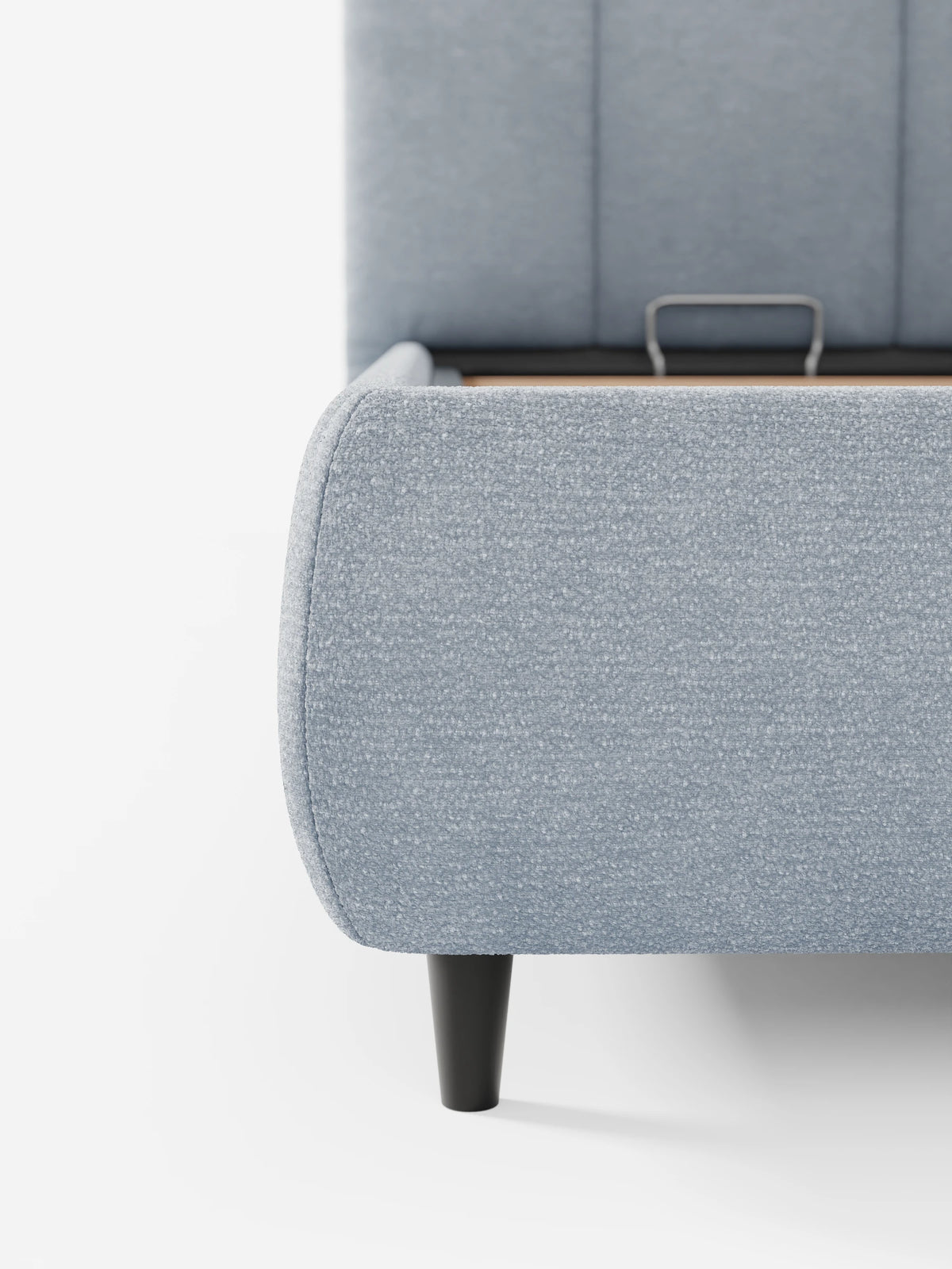 Un primo piano del divano letto ARKAM azzurro, imbottito, con vano contenitore e testiera a lavorazione verticale, caratterizzato da cuciture verticali sui pannelli, una maniglia di sollevamento in metallo e una base contenitiva parzialmente aperta sostenuta da gambe affusolate di colore scuro.