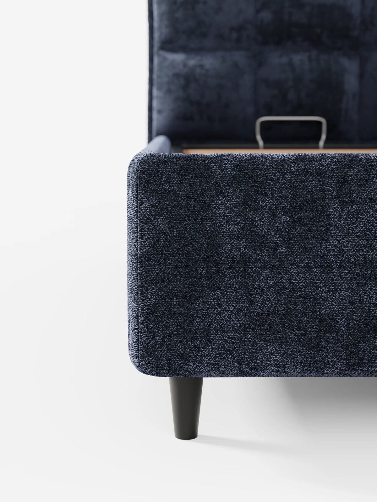 Un primo piano del letto imbottito AURO blu navy con vano contenitore e testiera alta capitonné in ciniglia Oeko-Tex®, caratterizzato da un tessuto testurizzato e da una gamba affusolata in legno scuro, raffigurato con la struttura sollevata per mostrare il vano contenitore interno.