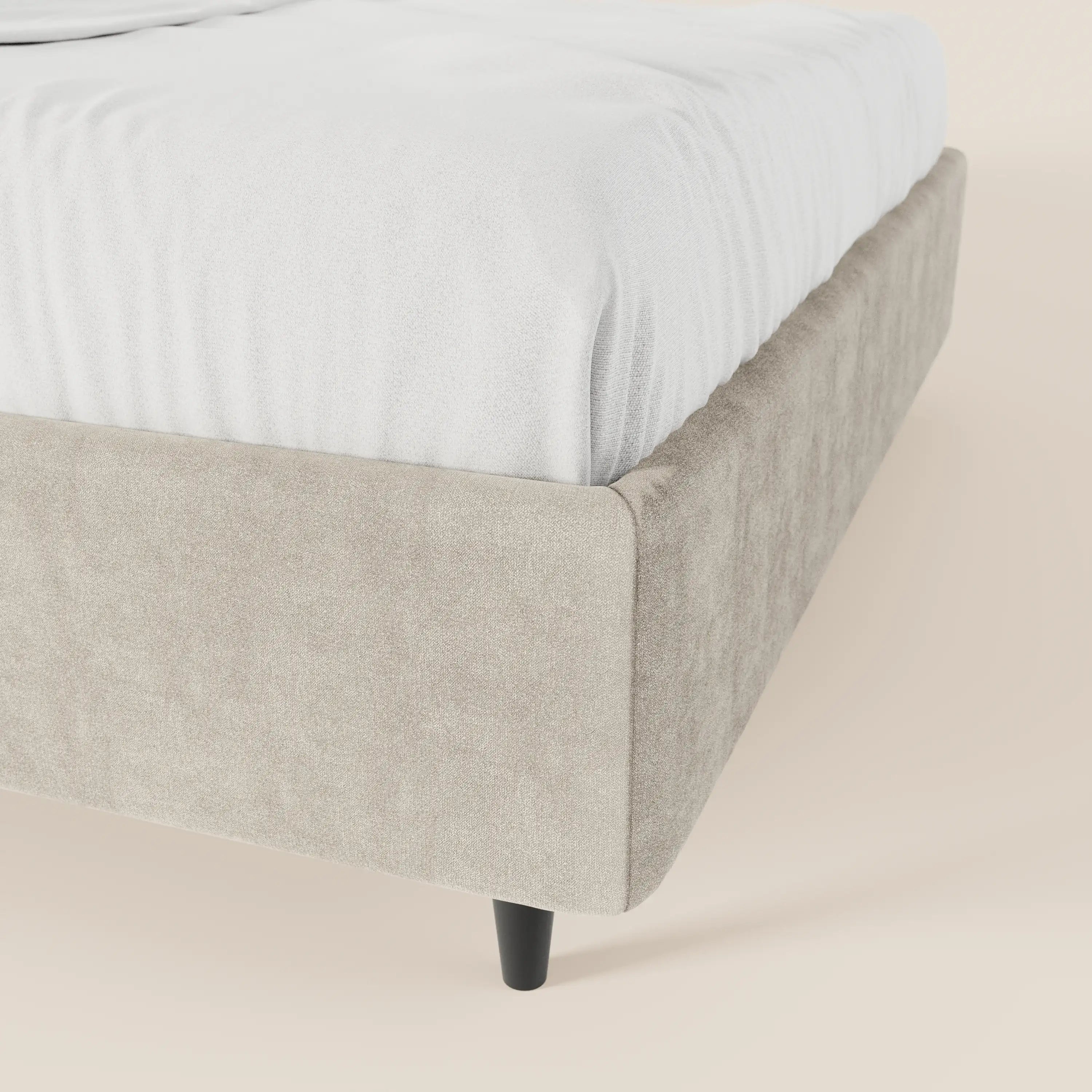 Primo piano del letto sommier imbottito ERON di Letti.store, che mostra il suo tessuto impermeabile beige certificato Oeko-Tex, la gamba affusolata in legno nero e il lenzuolo bianco ben teso. Lo sfondo presenta una superficie liscia e chiara.