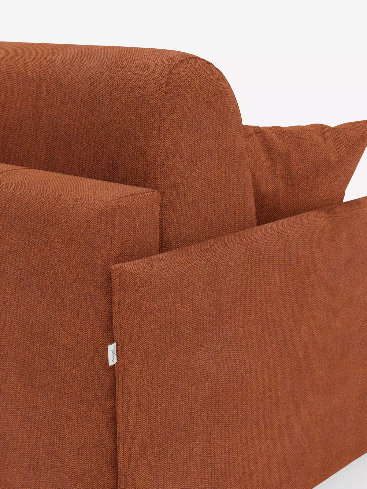 Un primo piano del divano letto matrimoniale sfoderabile Evans di Divani.Store — un divano letto compatto e moderno rivestito in tessuto T23 color ruggine e resistente alle macchie — che mostra il bracciolo, lo schienale, un cuscino coordinato e una piccola etichetta in tessuto bianco sul lato, su uno sfondo bianco a tinta unita.