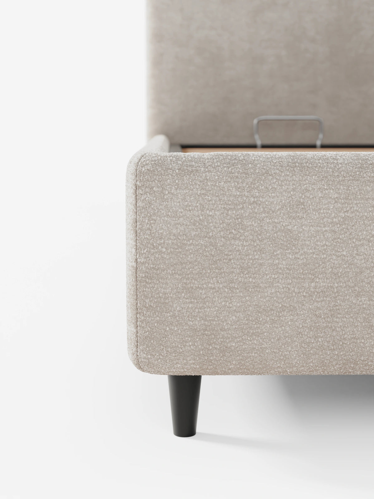 Un primo piano di un angolo del letto KAELOS minimal imbottito in ciniglia beige - con vano contenitore, piedini alti e testiera arrotondata, che mostra una gamba corta, scura e affusolata e una maniglia di sollevamento in metallo sulla base imbottita.