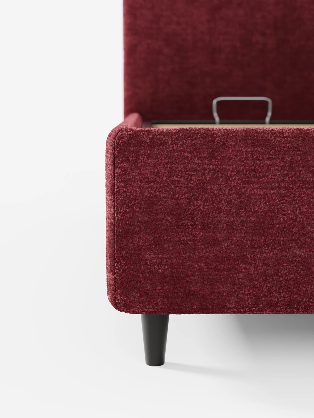 Un primo piano del letto KAELOS minimal imbottito in rosso intenso - con contenitore, gambe alte e testiera arrotondata in tessuto ciniglia, caratterizzato da un tessuto testurizzato, eleganti gambe nere e una maniglia di sollevamento in metallo sul telaio parzialmente aperto.