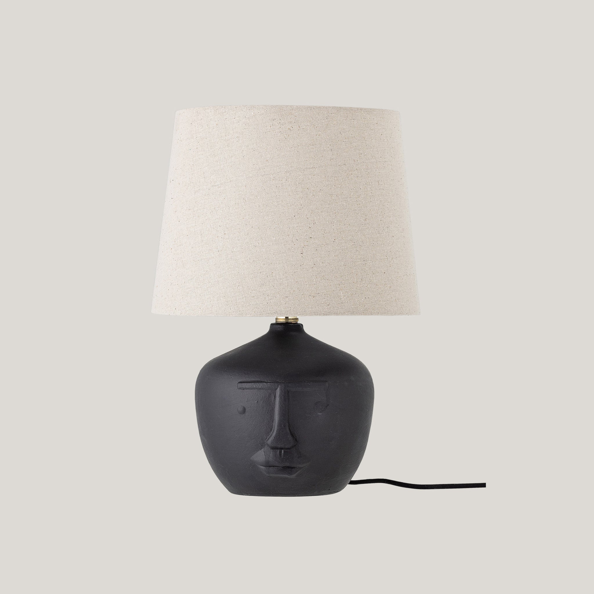 La lampada da tavolo Matheo di Divani.Store presenta un paralume in lino beige e una base in terracotta nera con un design stilizzato del volto, tra cui un naso alzato, occhi chiusi e una bocca sottile. Il suo cavo si estende verso destra su uno sfondo grigio chiaro.