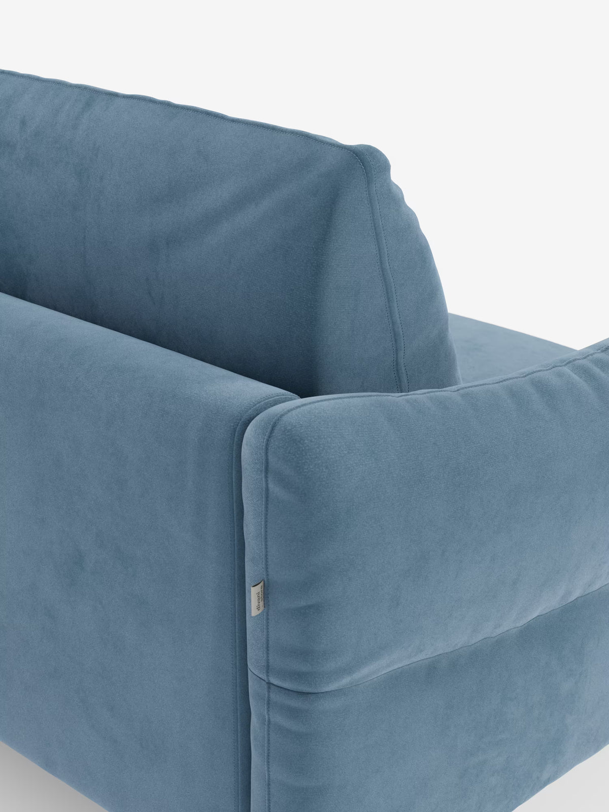 Un primo piano del moderno divano letto Dalia a 2, 3 e 4 posti con materasso H18 di Divani.Store, rivestito in tessuto di velluto T30 azzurro, facile da pulire. L'immagine mette in risalto i morbidi braccioli e lo schienale dalla consistenza vellutata e liscia, con cuciture a vista e una piccola etichetta in tessuto sul lato. Lo sfondo è bianco a tinta unita.