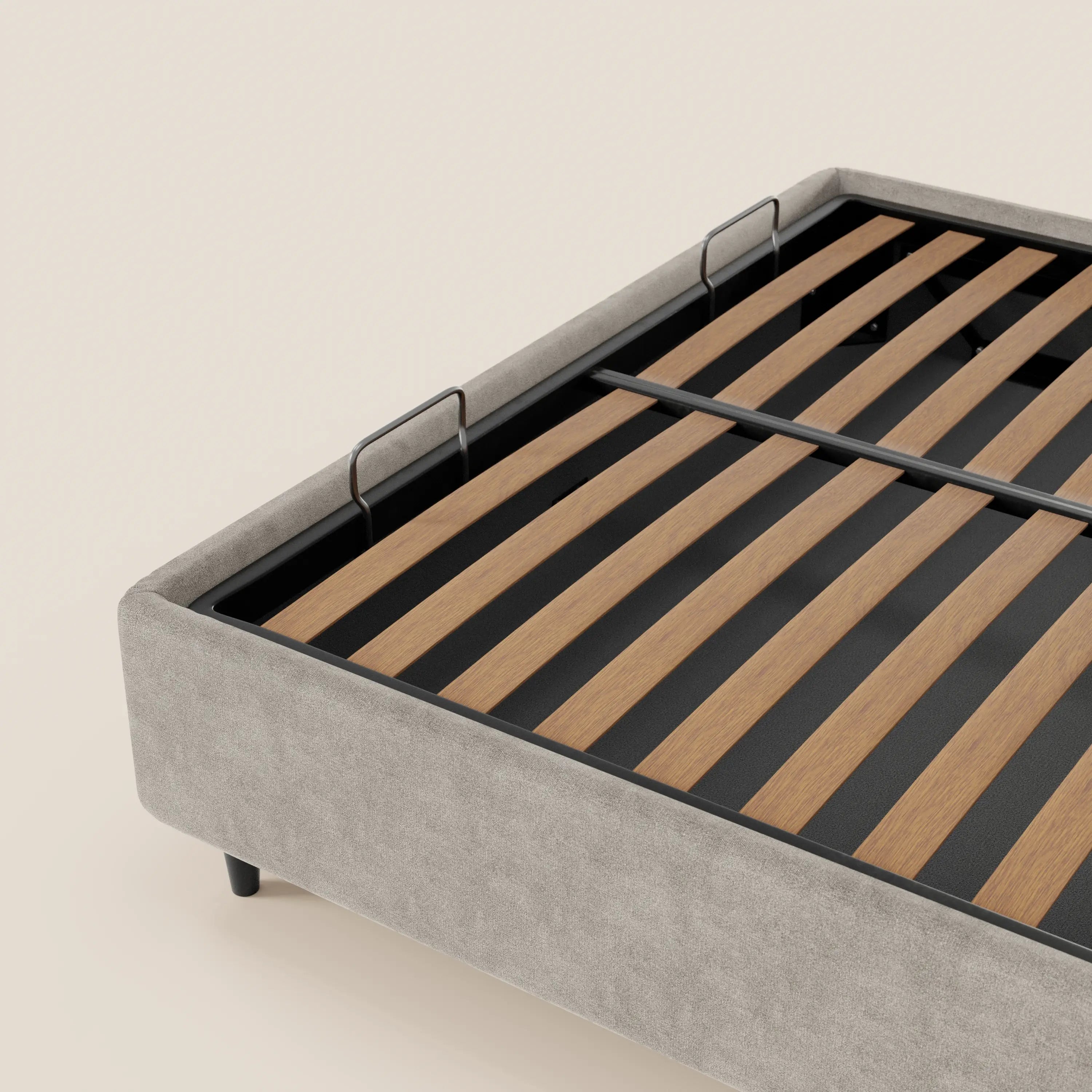 Un primo piano del letto sommier imbottito ERON di Letti.store, caratterizzato da una base in tessuto impermeabile Oeko-Tex grigio chiaro, bordo in metallo nero, doghe in legno, barre di sostegno in metallo e gambe corte nere su uno sfondo beige a tinta unita.