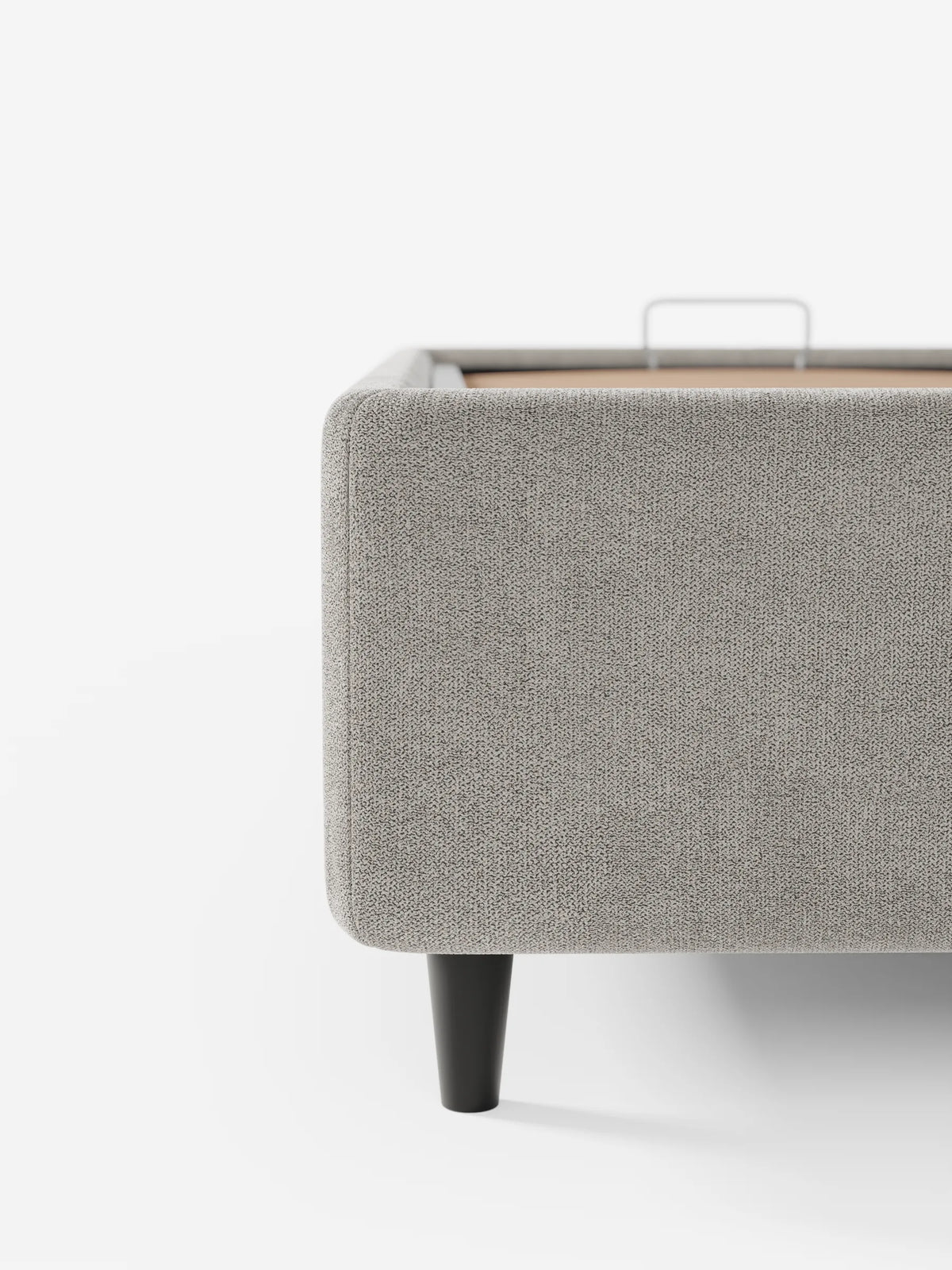 Primo piano di un letto sommier ERON grigio chiaro imbottito con vano contenitore in tessuto impermeabile certificato Oeko-Tex, dai bordi arrotondati, sostenuto da una gamba affusolata scura e dotato di una maniglia in metallo per l’accesso al vano contenitore.