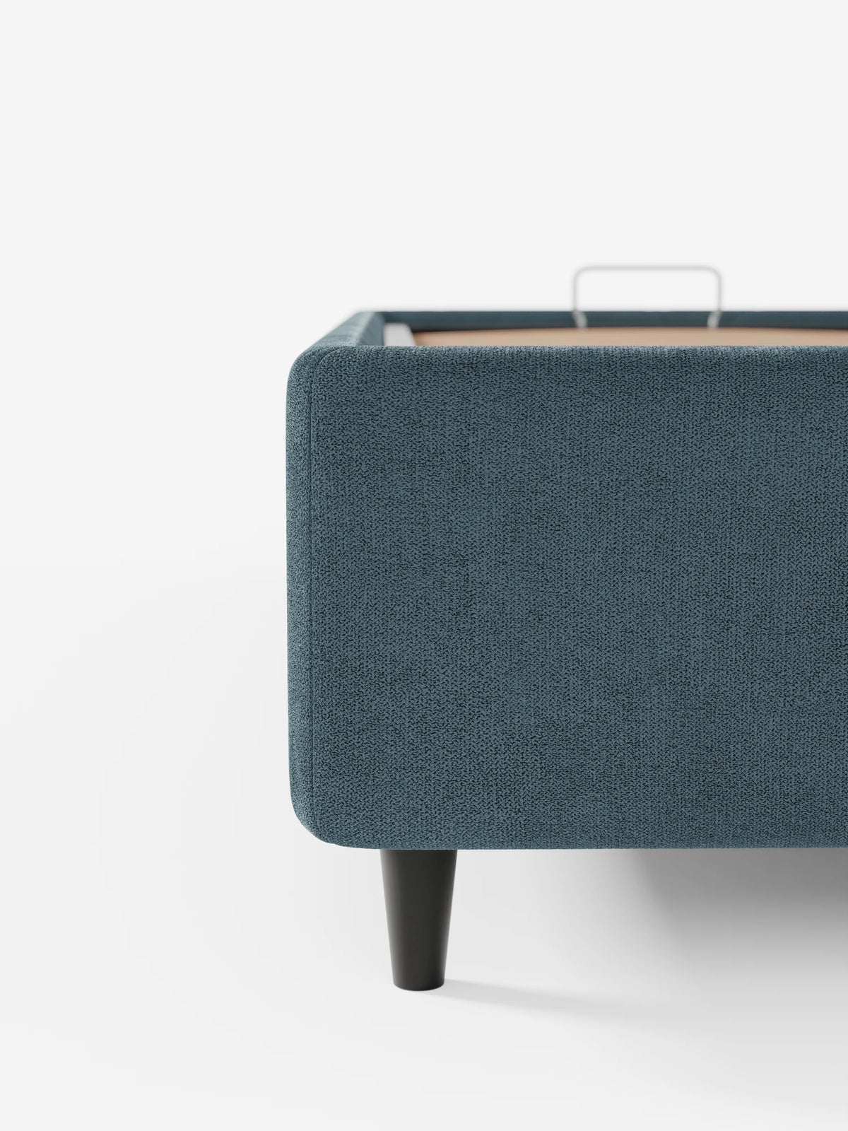 Un primo piano dell'angolo di un letto sommier imbottito ERON blu scuro con vano contenitore in tessuto impermeabile certificato Oeko-Tex, che mostra i bordi arrotondati, una gamba corta affusolata e una parte superiore aperta che lascia intravedere una struttura in legno chiaro e una maniglia in metallo.