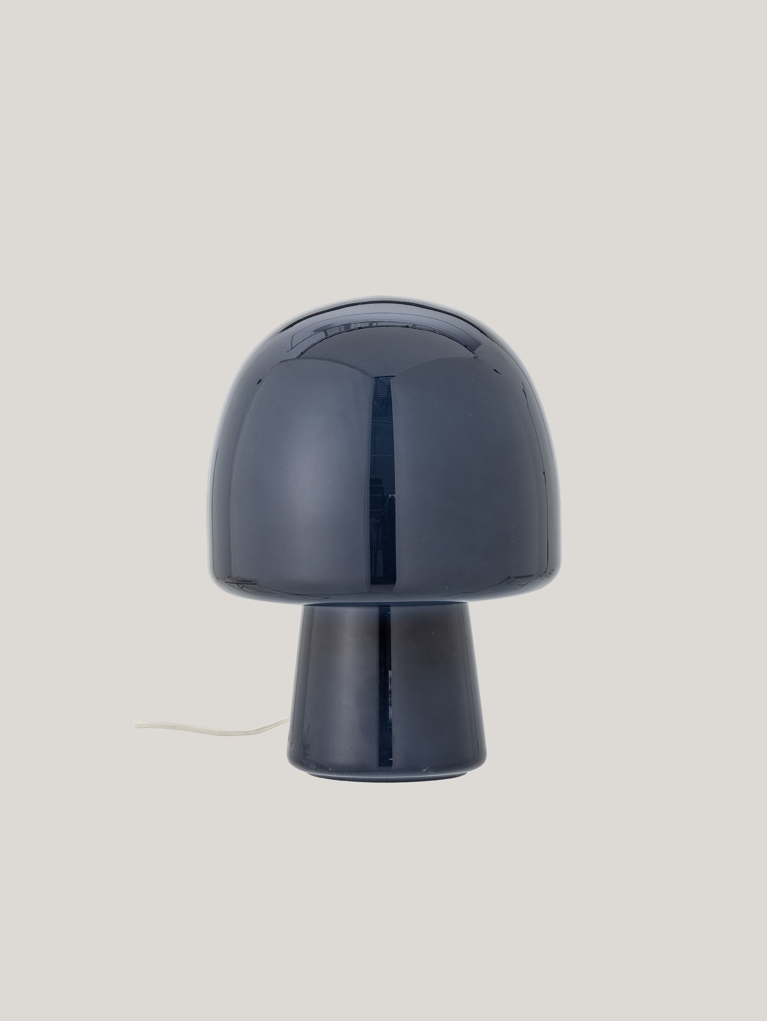 Un'elegante lampada da tavolo Paddy in una sofisticata tonalità blu, realizzata da Divani.Store, si staglia su uno sfondo semplice e grigio chiaro. La lampada presenta una cupola superiore che si trasforma senza soluzione di continuità in un'ampia base cilindrica che mette in mostra la maestria artigianale. Un cavo elettrico bianco emerge dal retro della base.