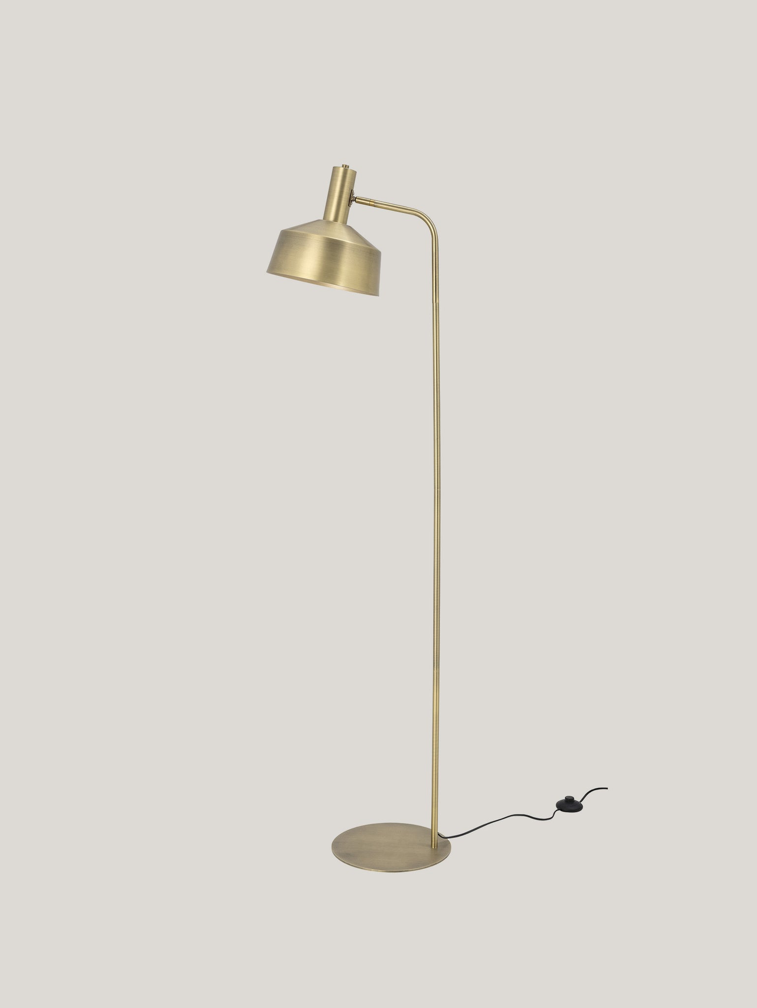 La lampada da terra Lissa in metallo colore ottone di Divani.Store presenta un design minimalista con base rotonda. Ha uno stelo sottile e leggermente curvo che sostiene un paralume regolabile a forma di cupola. Un cavo di alimentazione nero con interruttore a pedale è visibile sul pavimento, esaltandone l'eleganza classica sullo sfondo grigio chiaro.