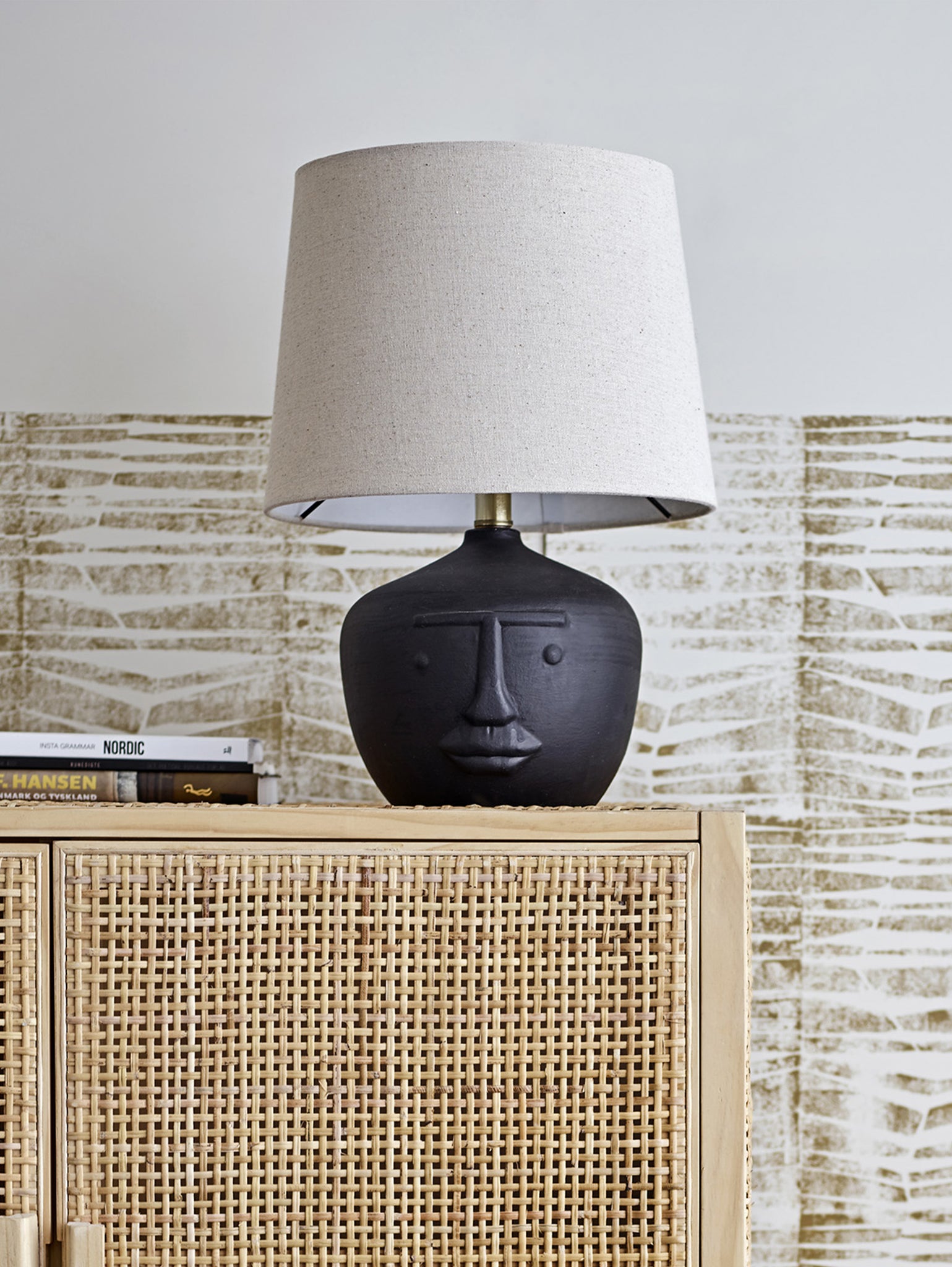 Una lampada da tavolo Matheo contemporanea di Divani.Store, con paralume in lino beige e base in terracotta scura con un design minimalista, è appoggiata su un mobile in rattan intrecciato. Il mobile in legno chiaro contiene libri e il motivo astratto testurizzato della parete esalta l'ambiente sofisticato.