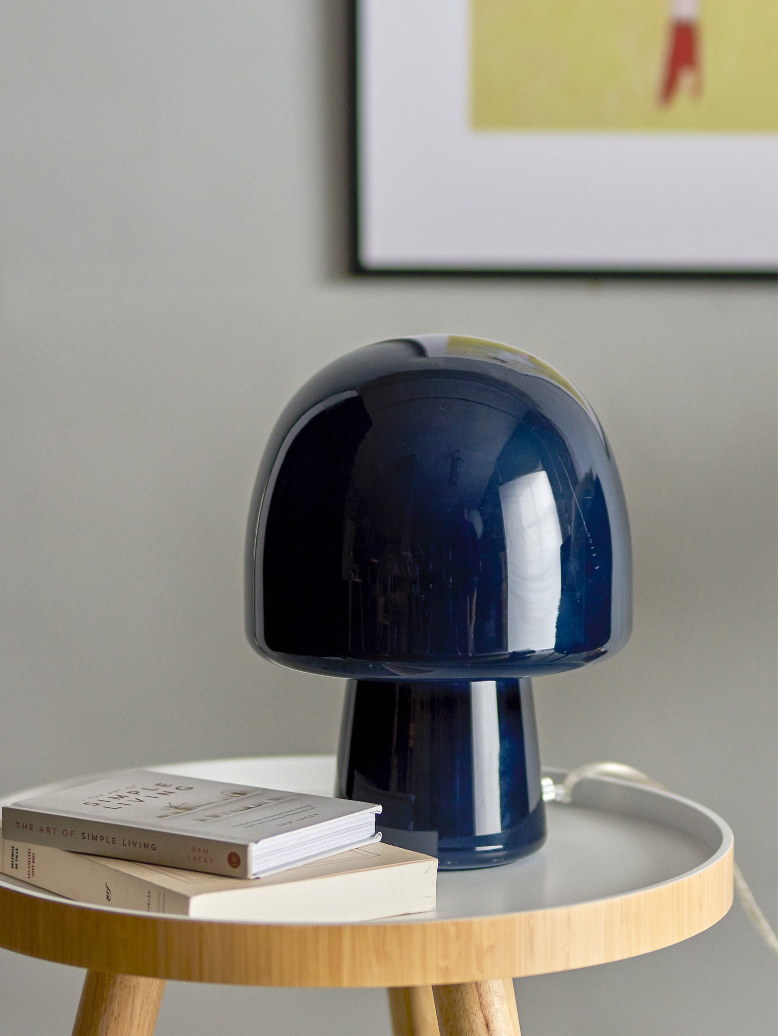 Una lampada da tavolo Paddy in vetro blu di Divani.Store è appoggiata su un tavolino rotondo in legno. Due libri, tra cui "The Art of Simple Living", sono impilati lì vicino. Sullo sfondo è presente un'opera d'arte incorniciata su una parete grigio chiaro.