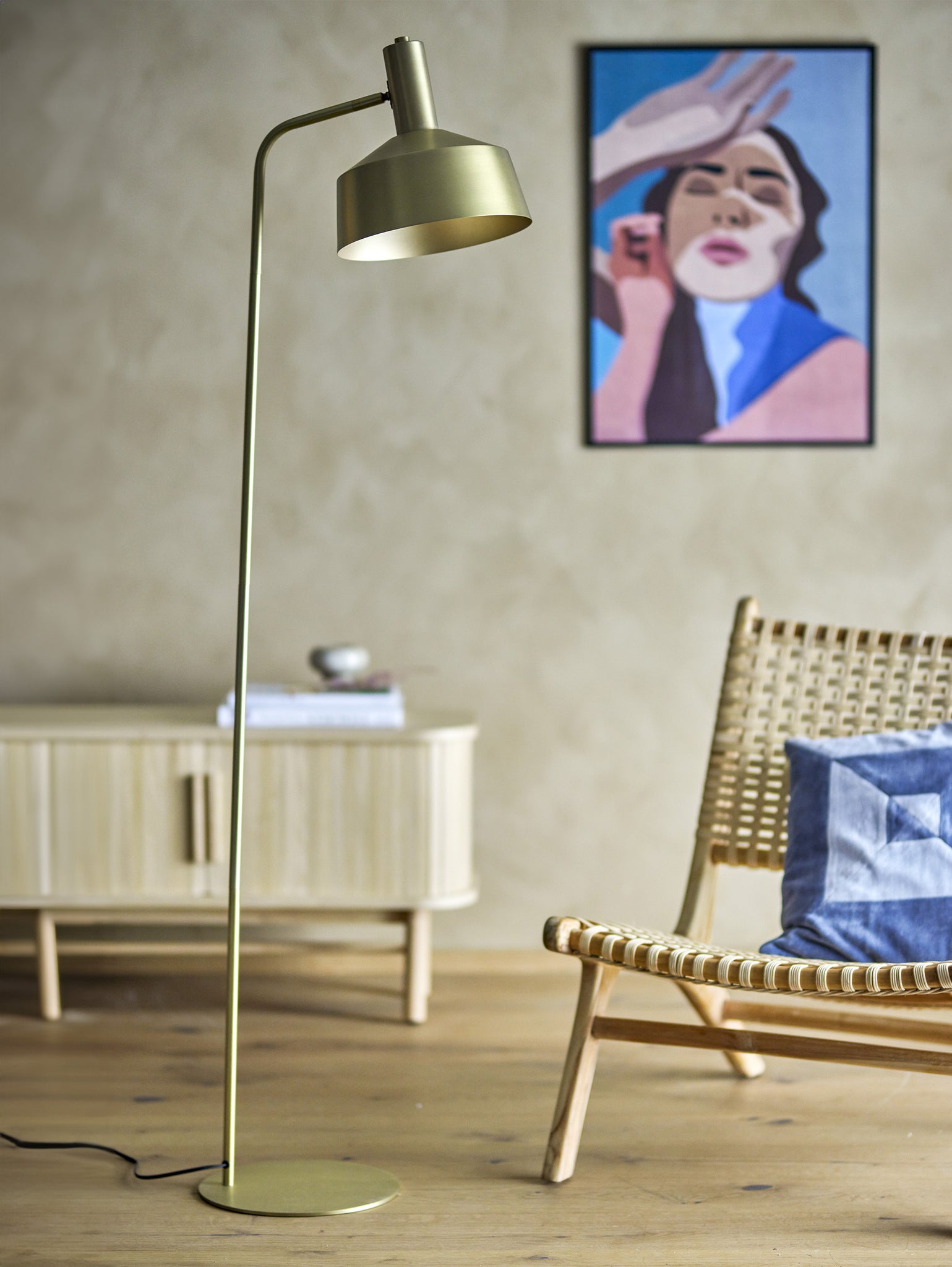 Un soggiorno moderno mette in mostra l'eleganza classica con una lampada da terra Lissa in metallo colore ottone di Divani.Store, una sedia intrecciata con un cuscino blu e una credenza in legno chiaro contro una parete beige. Un ritratto astratto colorato sopra aggiunge arte, mentre il pavimento in legno completa i toni caldi e neutri dello spazio.