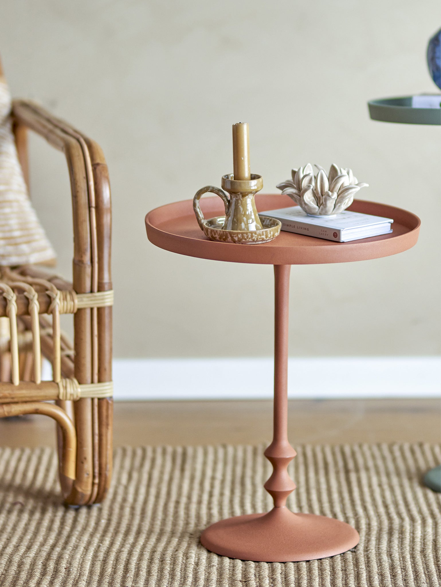 Un ambiente accogliente e minimalista presenta un tavolino da appoggio elegante Anjou in metallo arancio di Divani.Store, caratterizzato dalla sua forma piccola e rotonda e dalla base a stelo decorativa. Contiene una candela beige in un portacandele in vetro, un libro chiuso e un ornamento di loto in ceramica bianca. Posizionato su un tappeto intrecciato a righe, il tavolo è posizionato accanto a una sedia in vimini con cuscini beige.