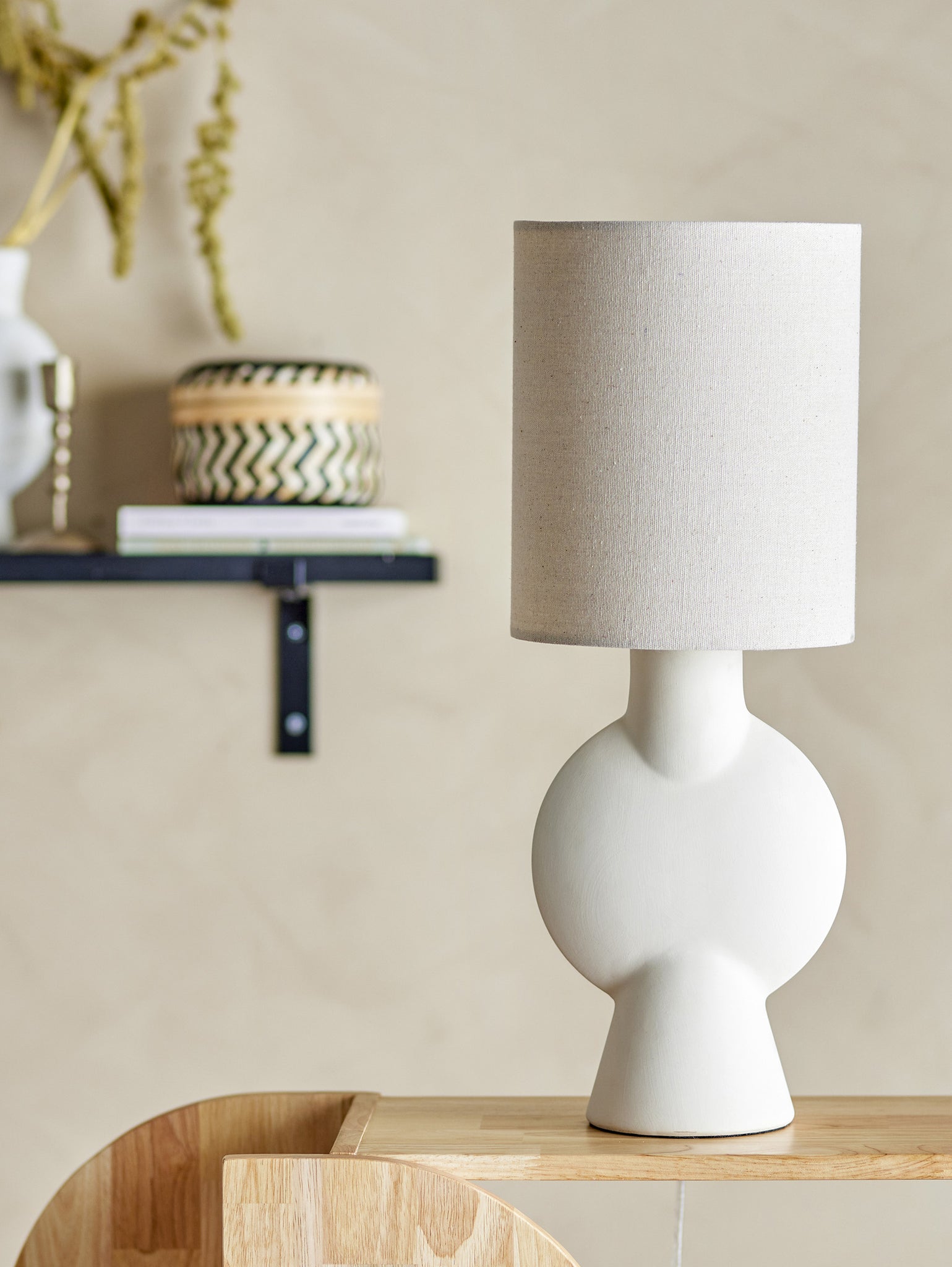 La lampada da tavolo Sergio di Divani.Store, con paralume beige lino e base in gres porcellanato finemente scolpita in bianco, è appoggiata su una superficie in legno. Sullo sfondo, una mensola nera espone un contenitore decorato, libri e un vaso pieno di fiori secchi contro una parete beige.