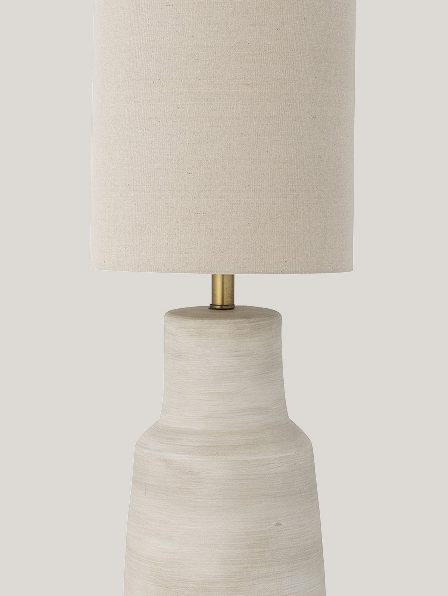 La lampada da tavolo Linetta di Divani.Store presenta un design minimalista, che mette in mostra un paralume cilindrico in tessuto beige abbinato a una base in ceramica testurizzata in gres porcellanato color avorio. Un elegante connettore in ottone unisce senza soluzione di continuità il paralume e la base sullo sfondo chiaro e neutro.