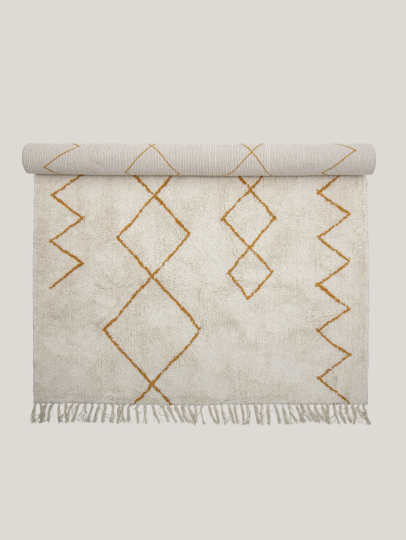 Il prodotto denominato "Huso tappeto a fantasia geometrica in cotone 200x140 cm" di Divani.Store presenta un sofisticato motivo a rombi in giallo senape su uno sfondo beige. Il tappeto è leggermente arrotolato nella parte superiore, esponendo il retro a strisce, e nappe a frange impreziosiscono il bordo inferiore sullo sfondo chiaro e solido.