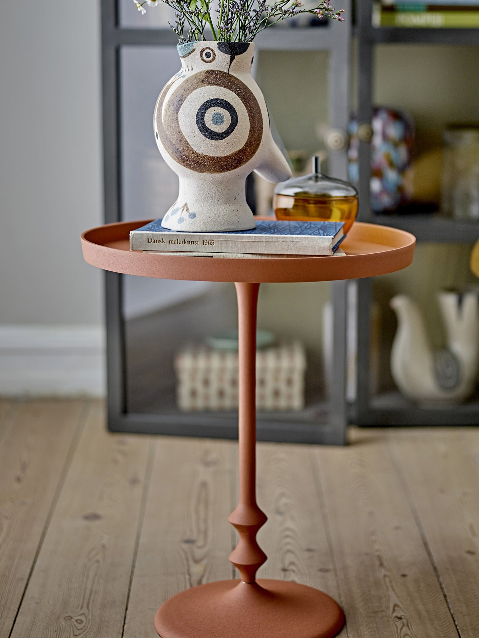 L'Anjou tavolino da appoggio elegante in metallo arancio di Divani.Store presenta un vaso scultoreo decorato con motivi circolari e fiori sul suo design moderno. Sotto il vaso ci sono due libri, mentre un mobiletto con oggetti decorativi è sullo sfondo. Questo elegante tavolo poggia con grazia su un pavimento in legno.