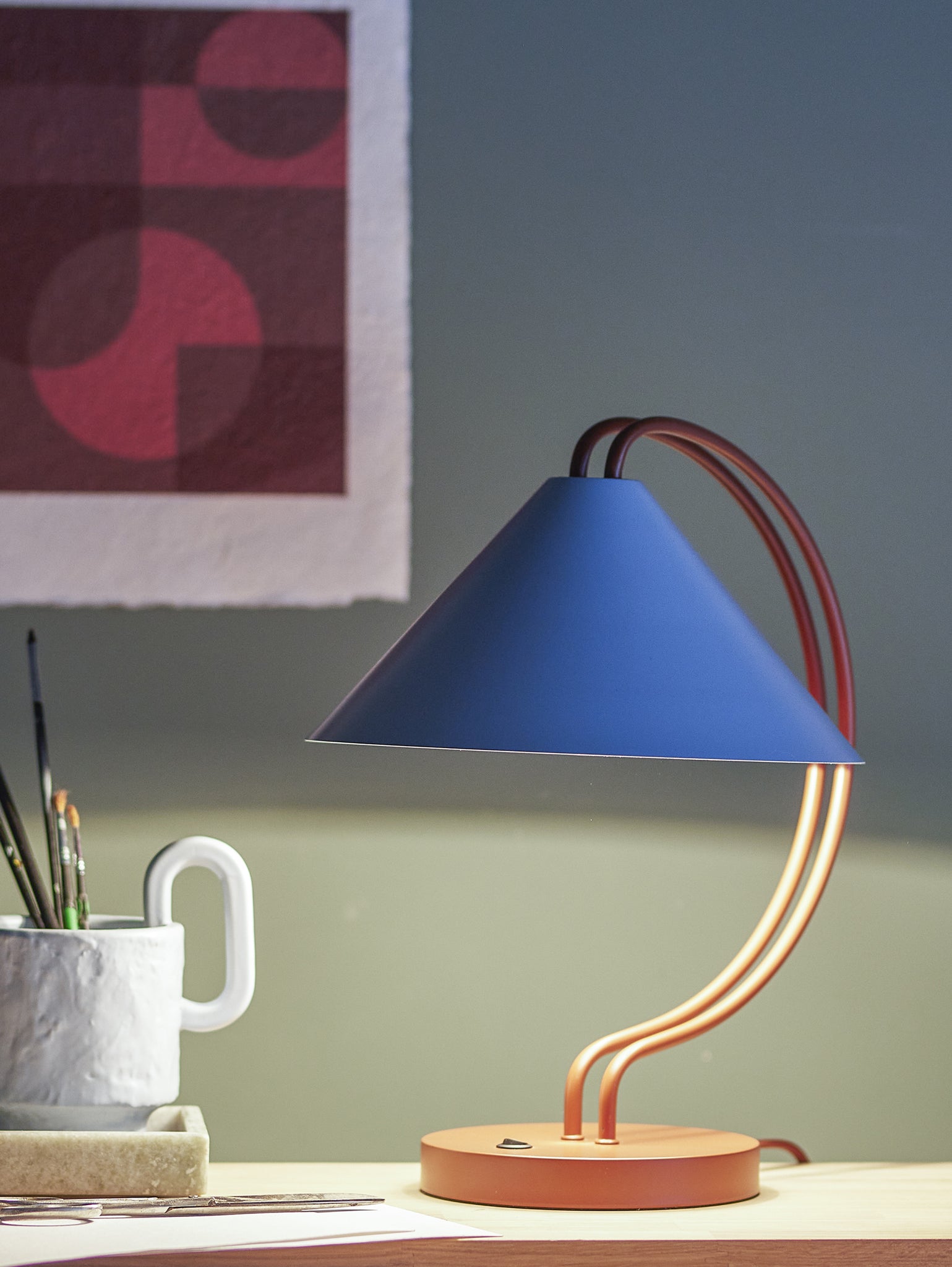 Un'elegante lampada da tavolo Janya di Divani.Store presenta un paralume conico blu e un supporto rosso curvo, appoggiato su una scrivania in legno con distinti accenti grafici nel suo design. Anche la base rotonda è rossa. Nelle vicinanze, una tazza bianca contiene dei pennelli, mentre un'opera d'arte con motivi geometrici in tonalità rosse adorna la parete dietro, su uno sfondo verde tenue.